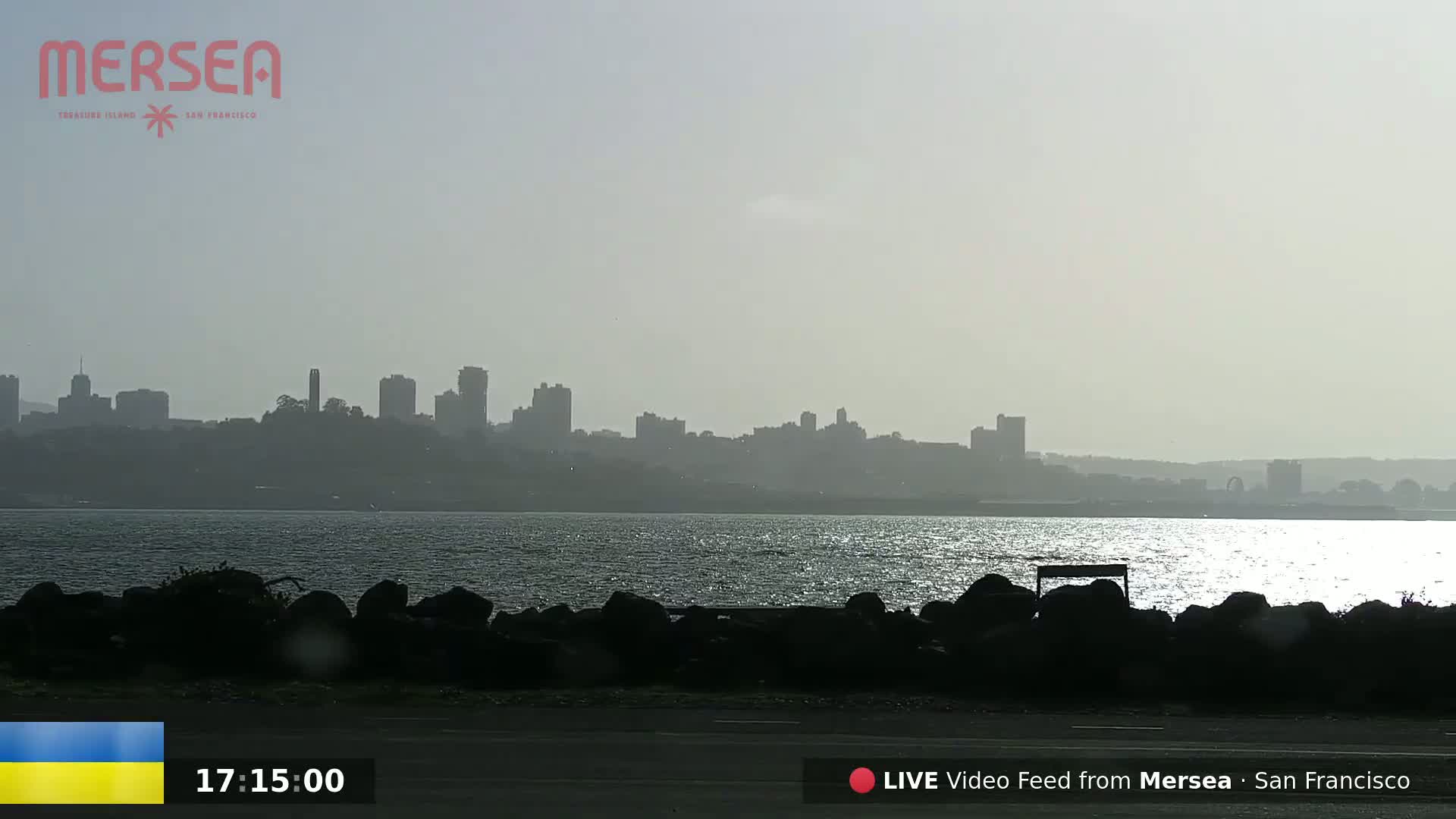 A hazy cityscape is visible across a body of water from a shoreline with dark rocks and a bench.