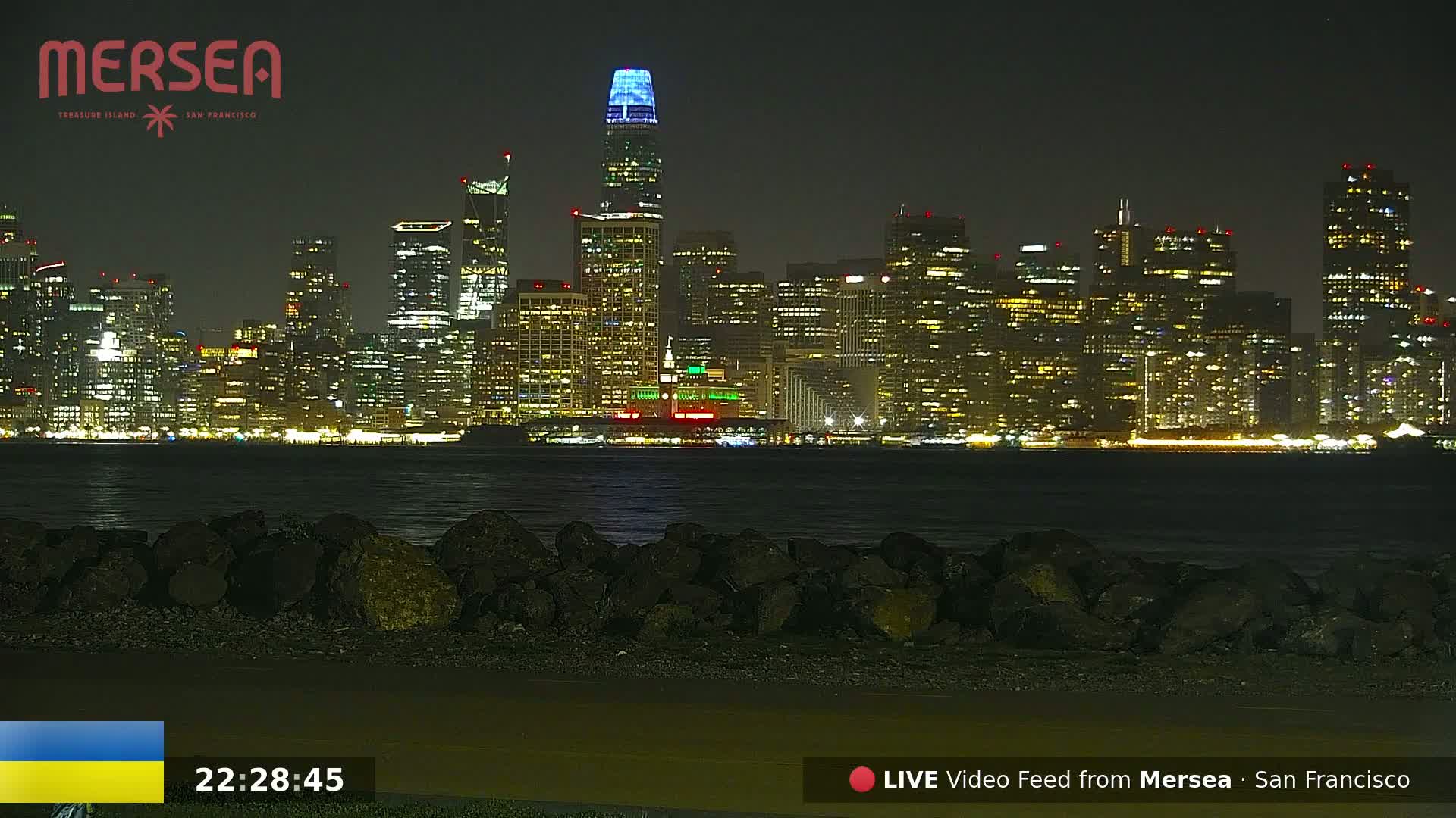 A nighttime view of a city skyline across a body of water, with dark rocks in the foreground.