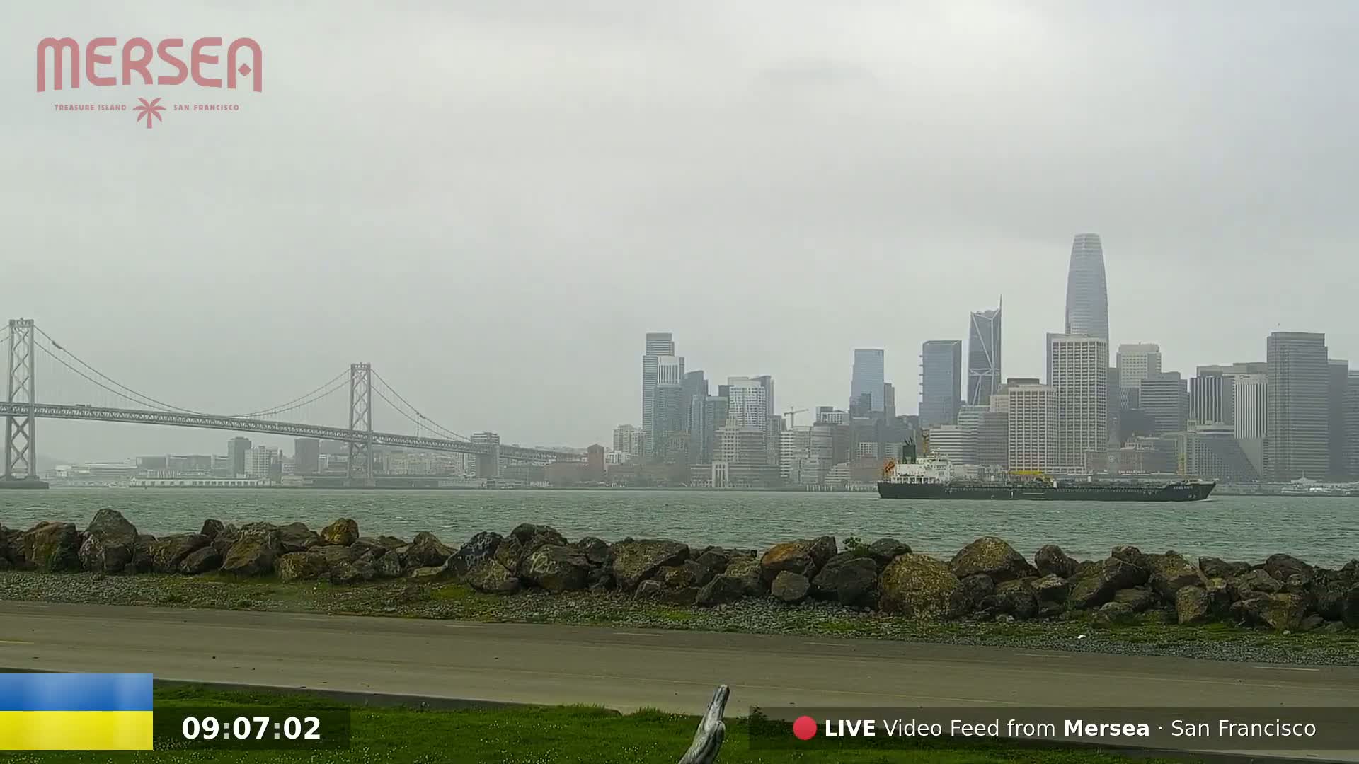 A large cargo ship is in the water in front of a hazy San Francisco skyline, with a bridge visible in the distance.