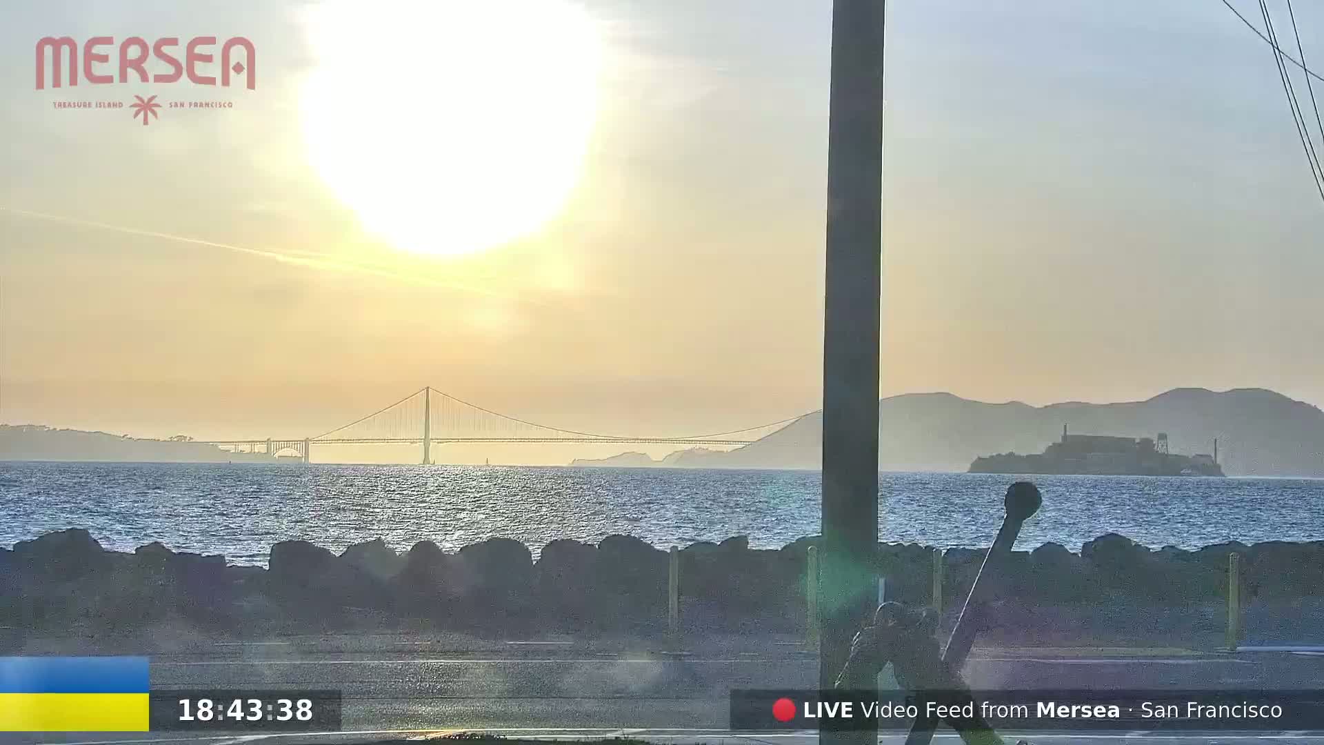 A sunlit sunset view shows a suspension bridge and Alcatraz Island across a calm body of water, with a rocky shoreline in the foreground.