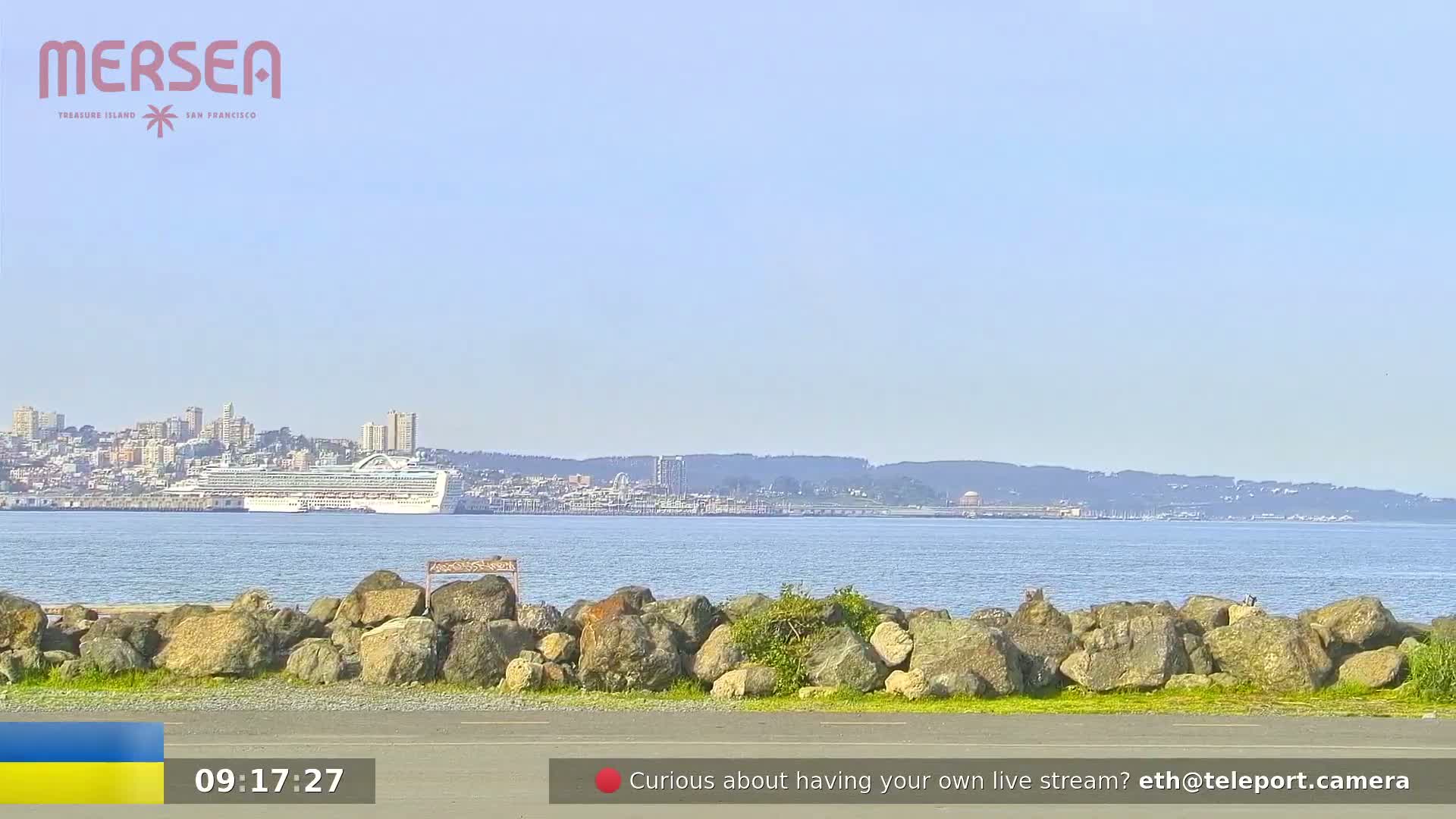 A large cruise ship is anchored in a calm bay, with a city skyline visible in the background on a clear, sunny day.