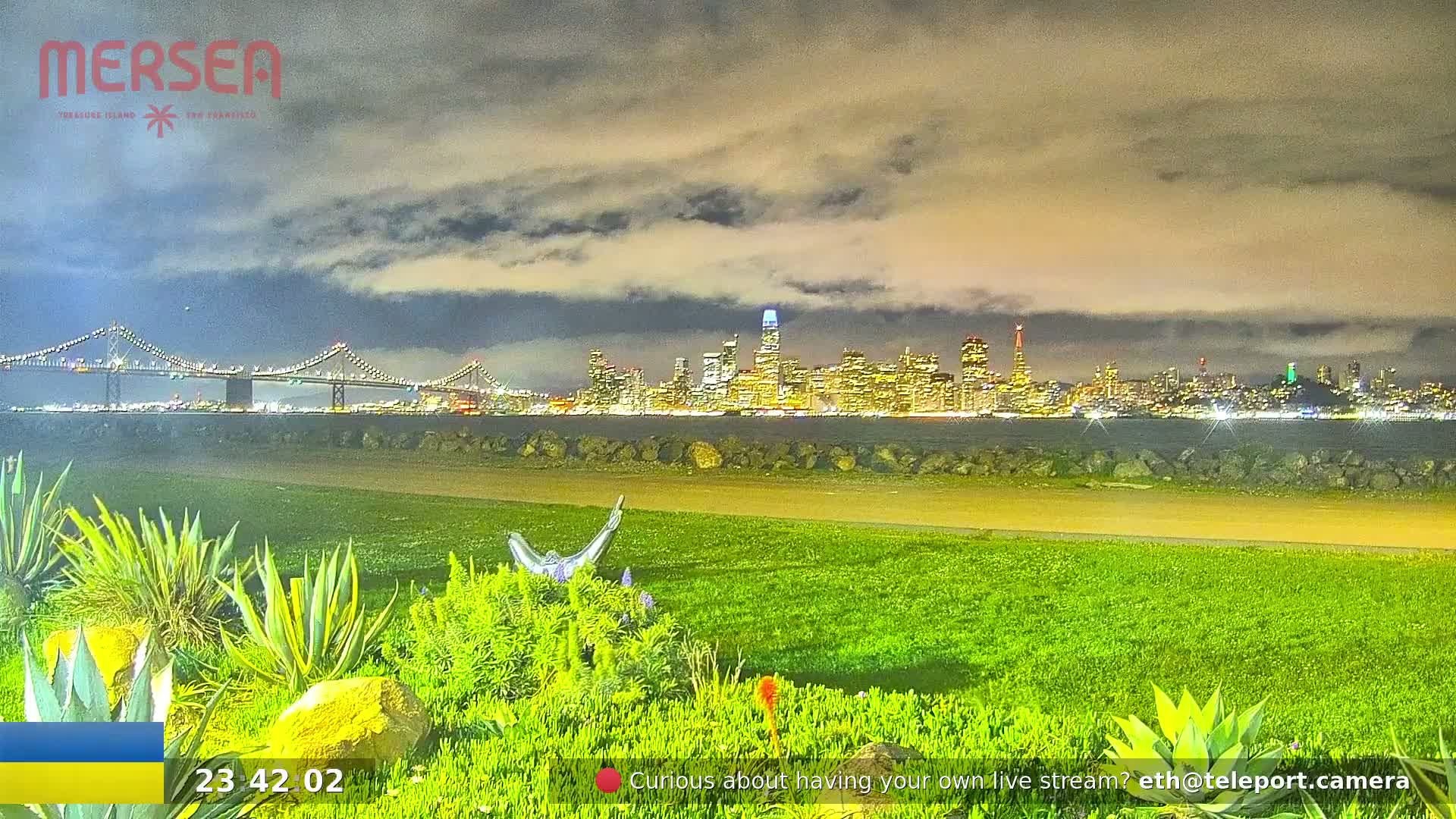 A nighttime view of a city skyline across a body of water, under a partly cloudy sky, with a bridge in the distance and landscaping in the foreground.