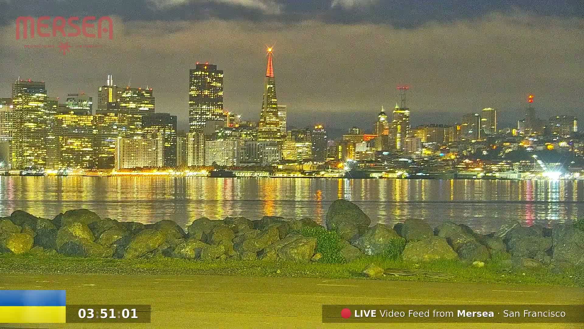 A nighttime view of a city skyline brightly lit across a calm body of water, with a rocky shoreline in the foreground under a cloudy sky.