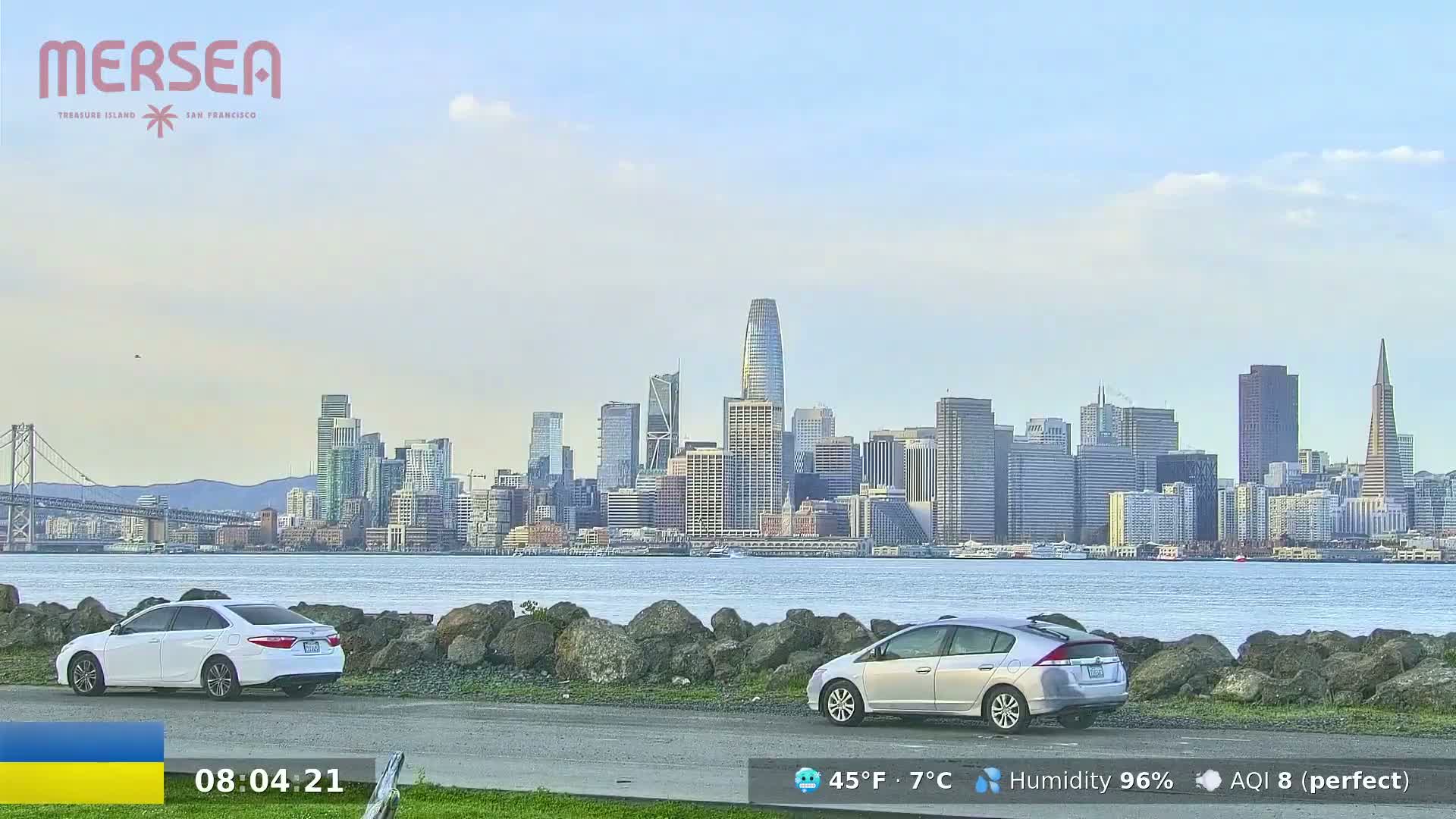 Two cars are parked near a body of water with a city skyline in the background under a mostly clear sky.