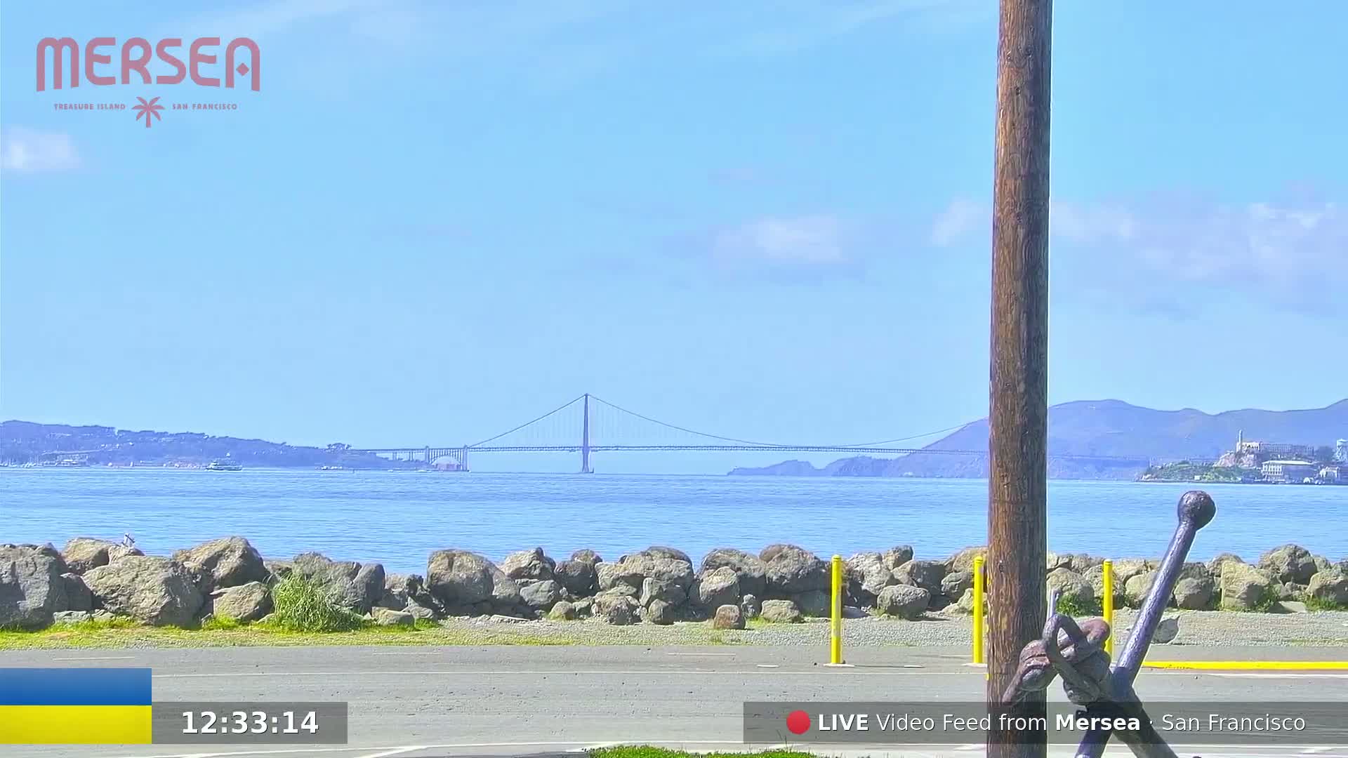 A suspension bridge is visible in the distance across a calm body of water, under a mostly clear blue sky with a few scattered clouds, from a waterfront perspective with large rocks and a portion of an anchor in the foreground.