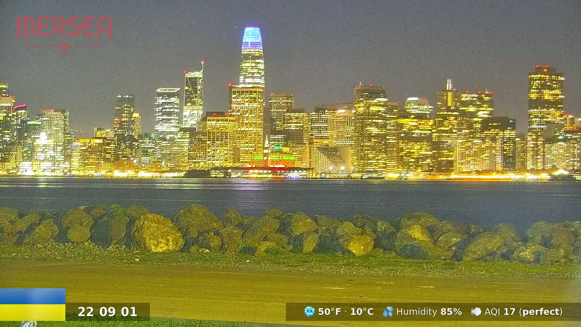 A brightly lit city skyline across a dark body of water, viewed from a rocky shoreline at night, under a humid sky.