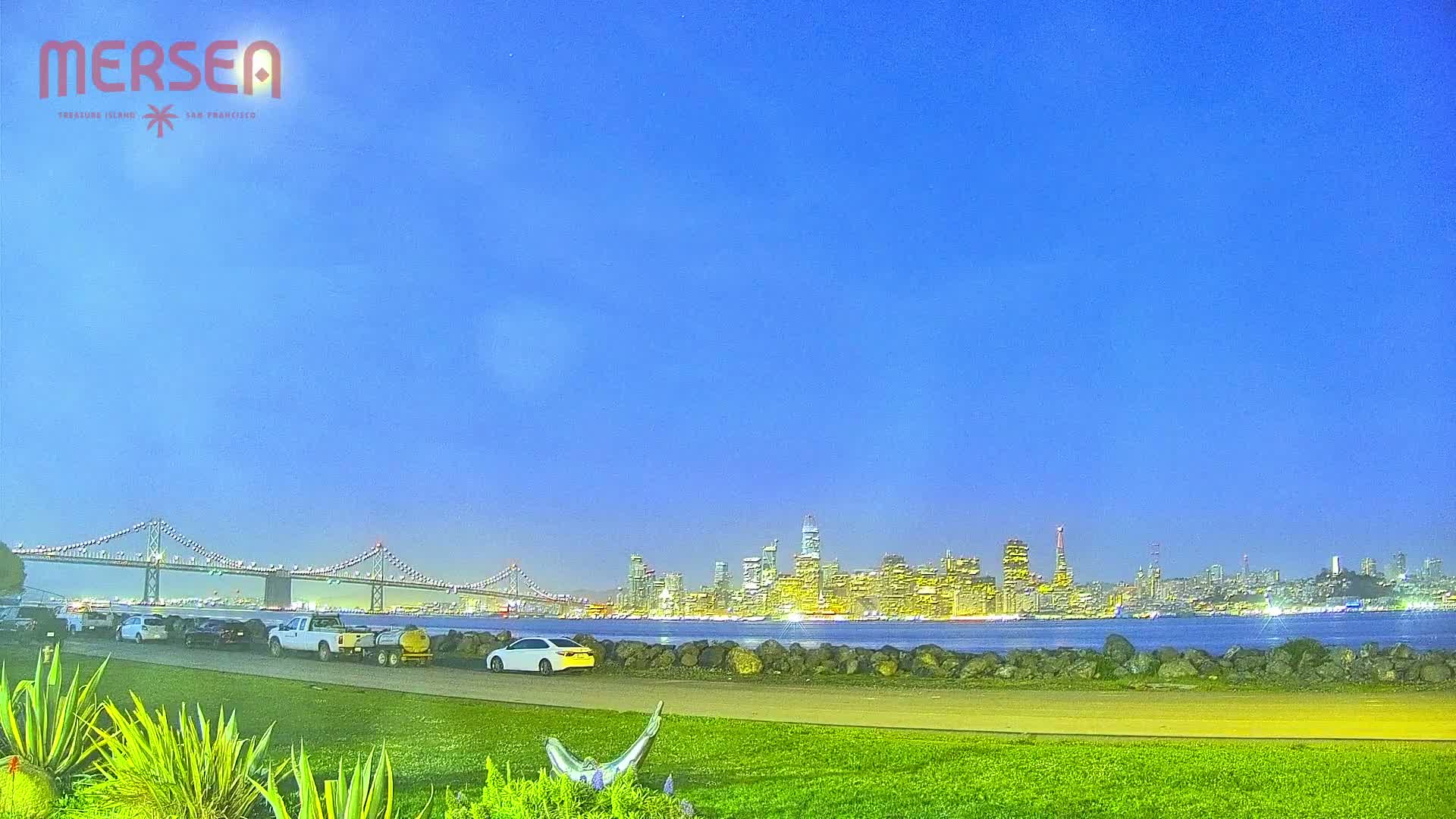 Under a clear twilight sky, a grassy area overlooks a body of water with a cityscape and a bridge in the distance.