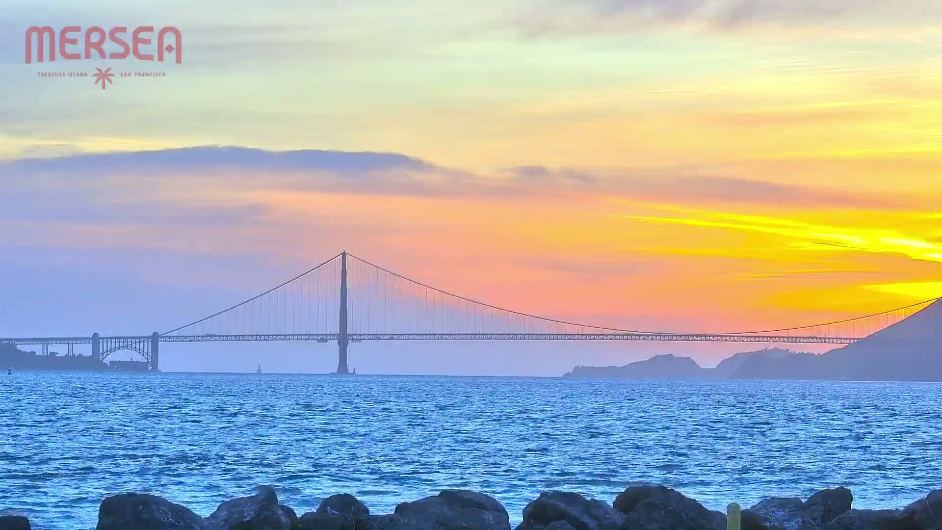 A suspension bridge spans a body of water under a colorful sunset sky.