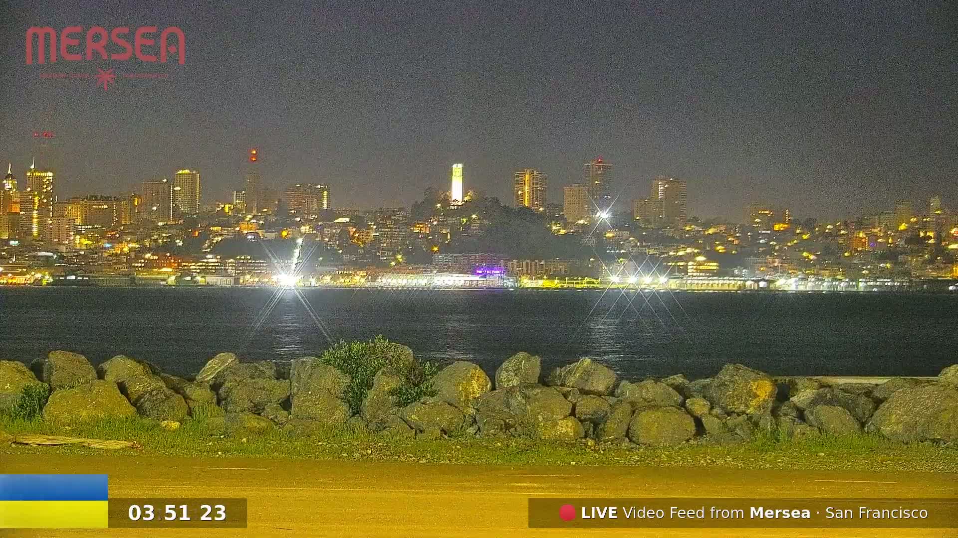 A nighttime view of a city skyline across a body of water, with a rocky shoreline in the foreground.