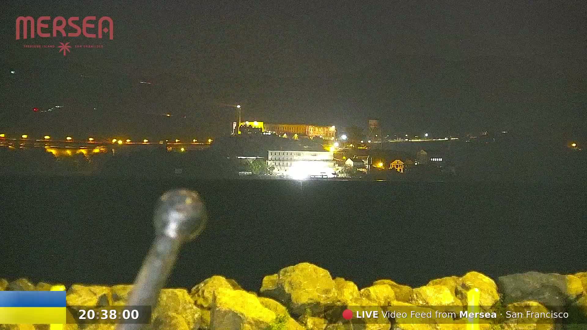 A nighttime long shot shows a brightly lit building complex across a dark body of water, with large rocks in the foreground.