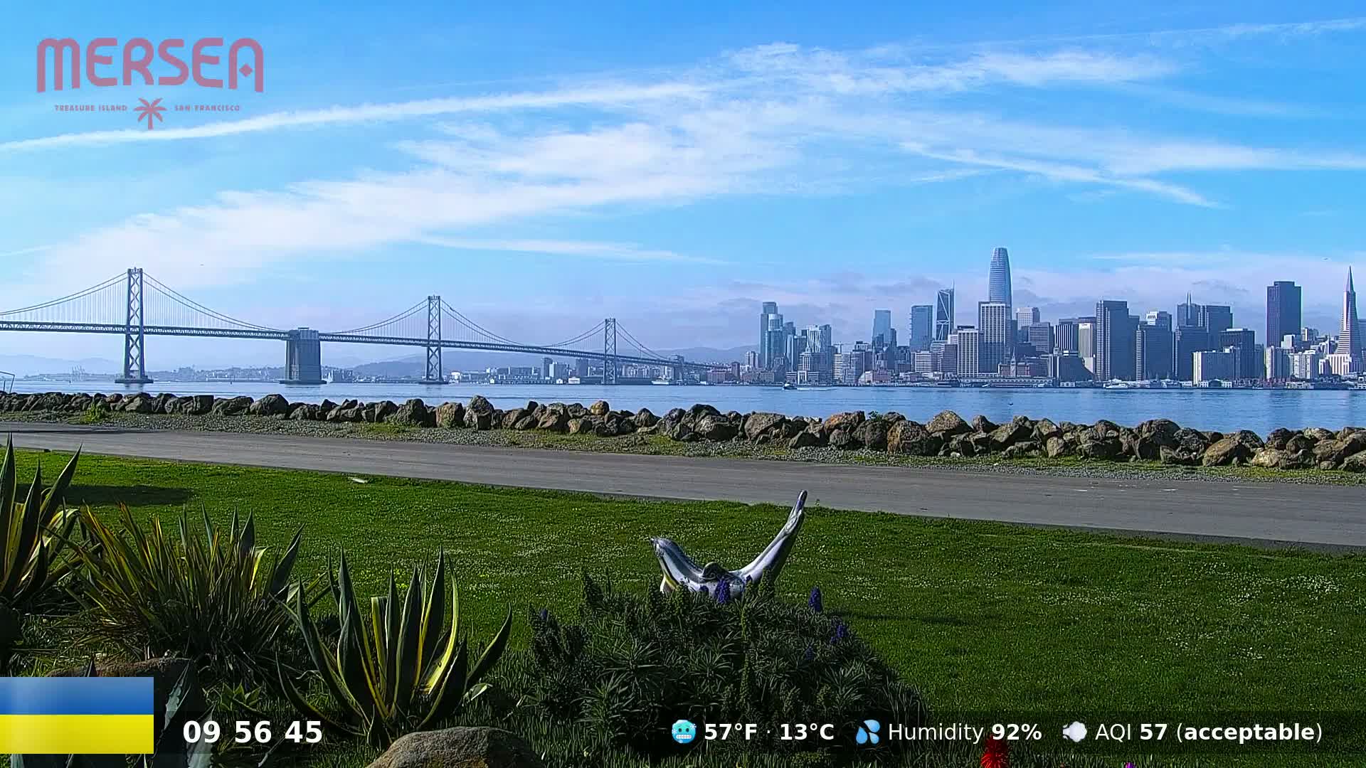 A grassy park with a metal sculpture overlooks a bay, a suspension bridge, and the San Francisco skyline under a mostly clear blue sky.