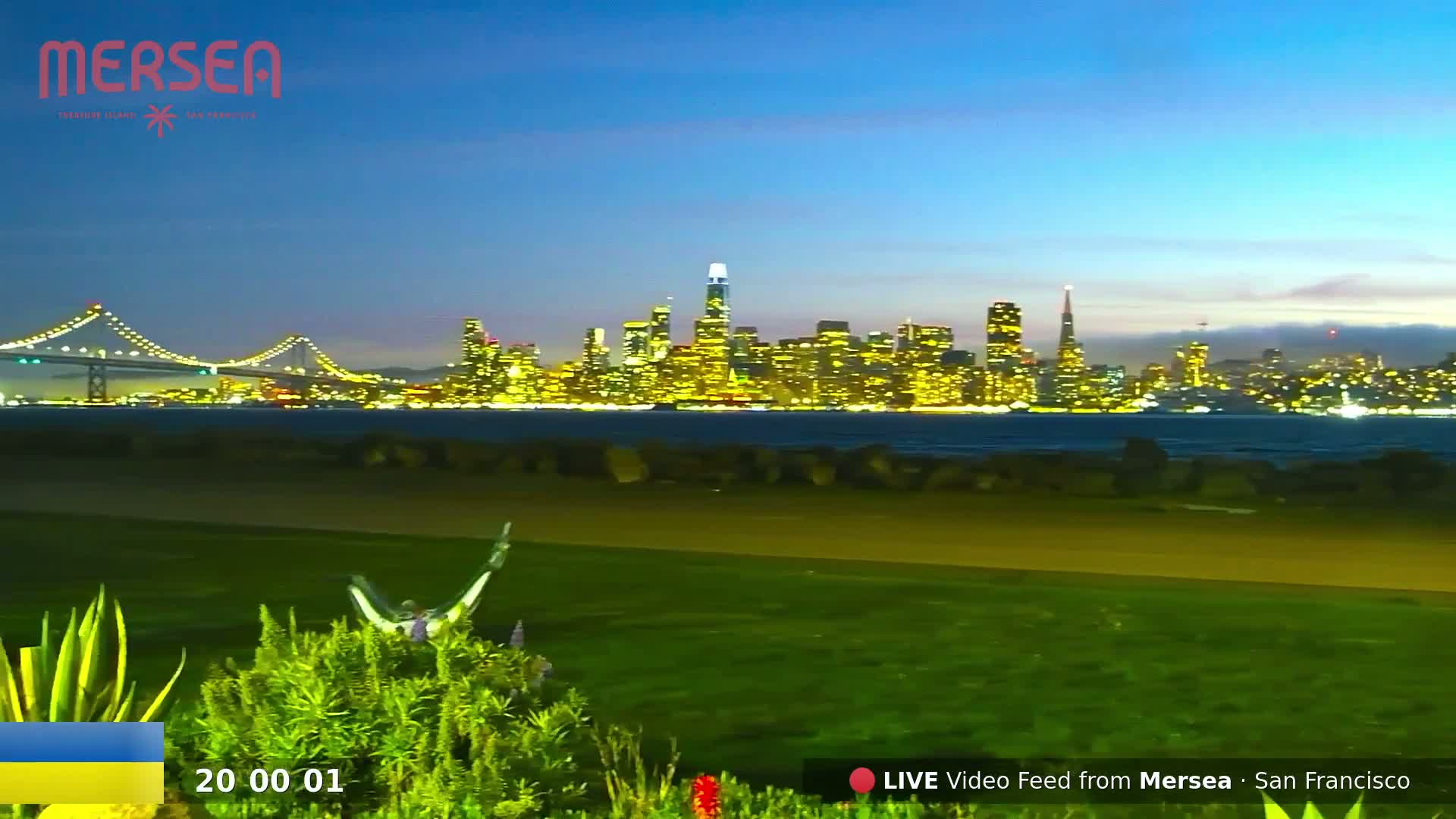 A brightly lit city skyline across a body of water at dusk, viewed from a grassy area with a person in a hammock visible in the foreground.