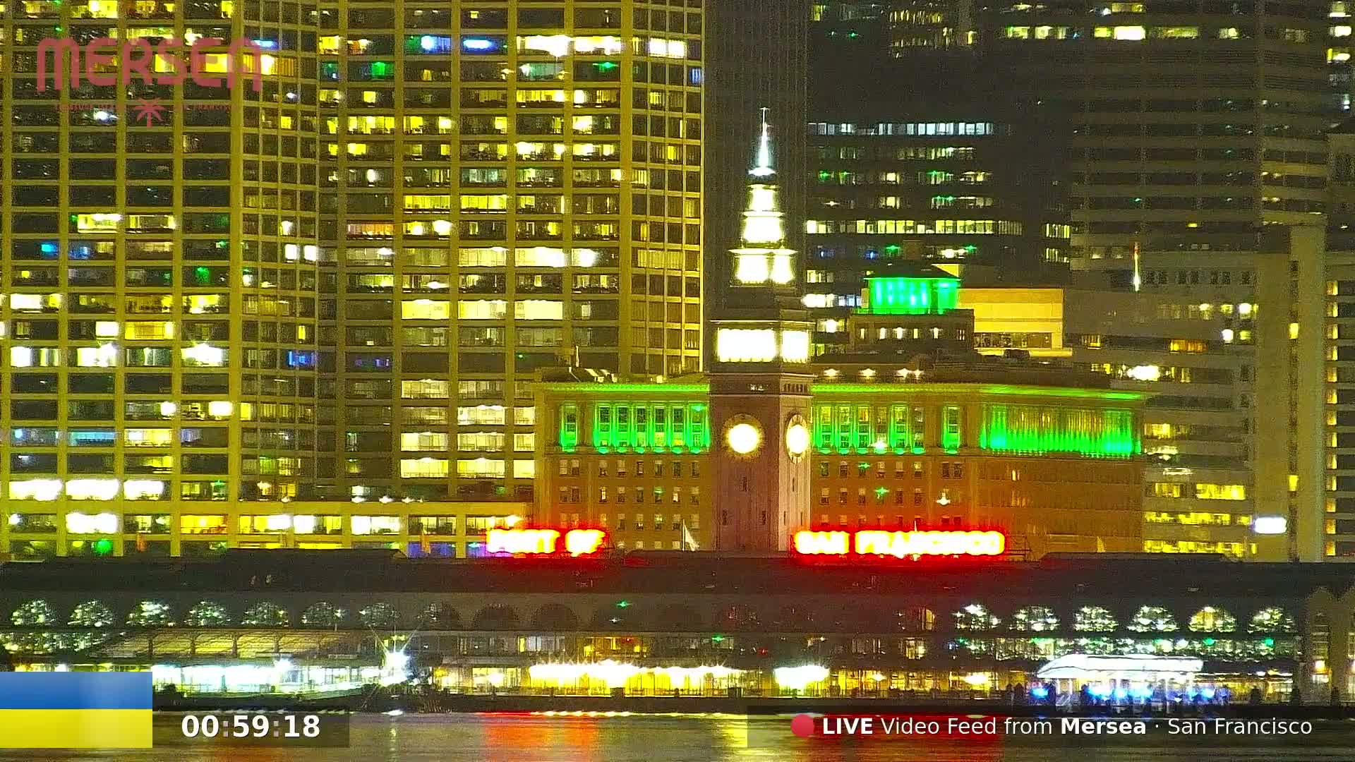 A nighttime cityscape of illuminated buildings near a body of water, with some buildings brightly lit in green and yellow.