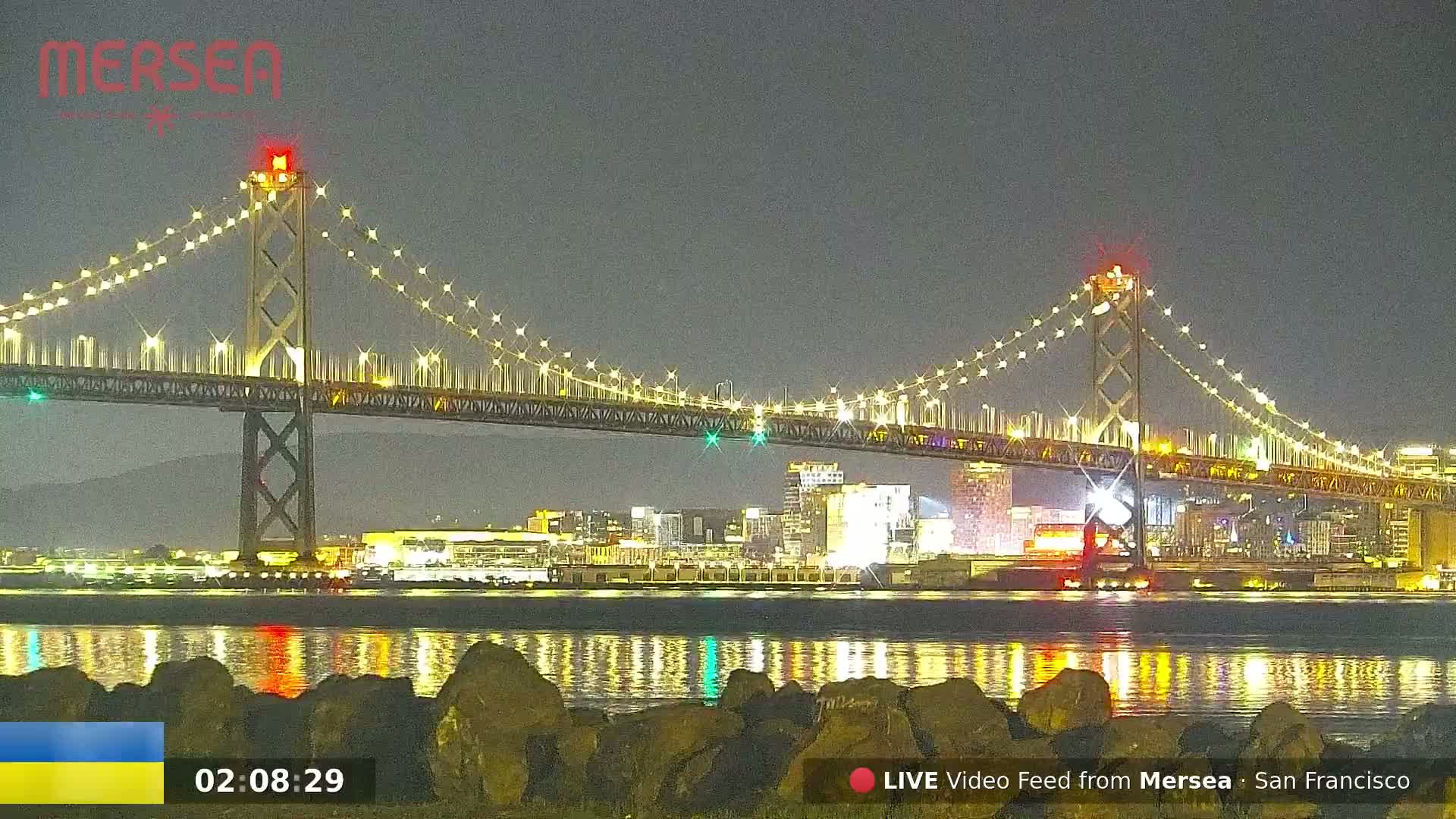 A brightly lit suspension bridge spans a dark body of water at night, with city lights reflected in the calm water.