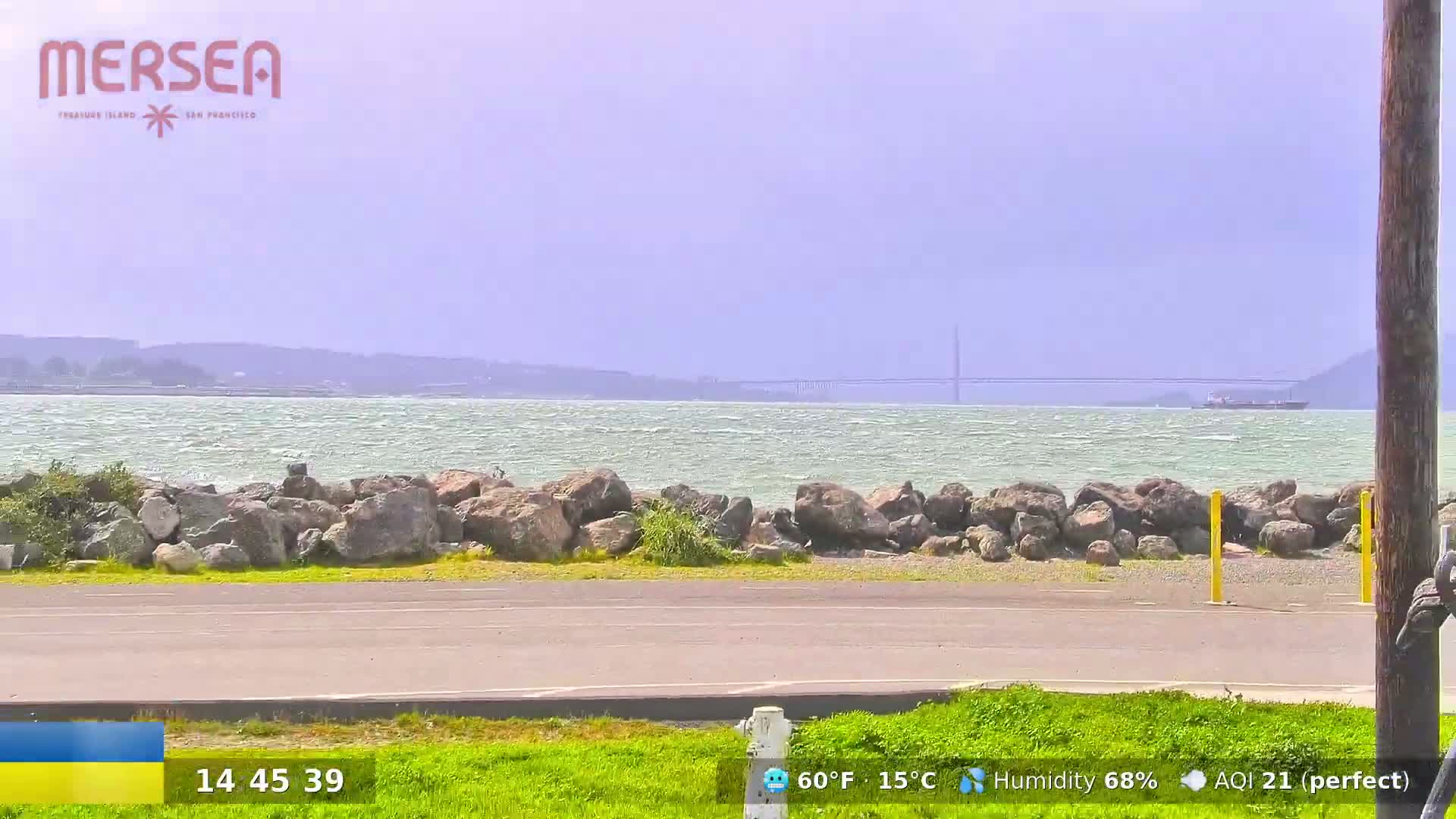 A partly cloudy day shows a rocky shoreline with a paved road next to a grassy area, overlooking a choppy body of water and a distant bridge.