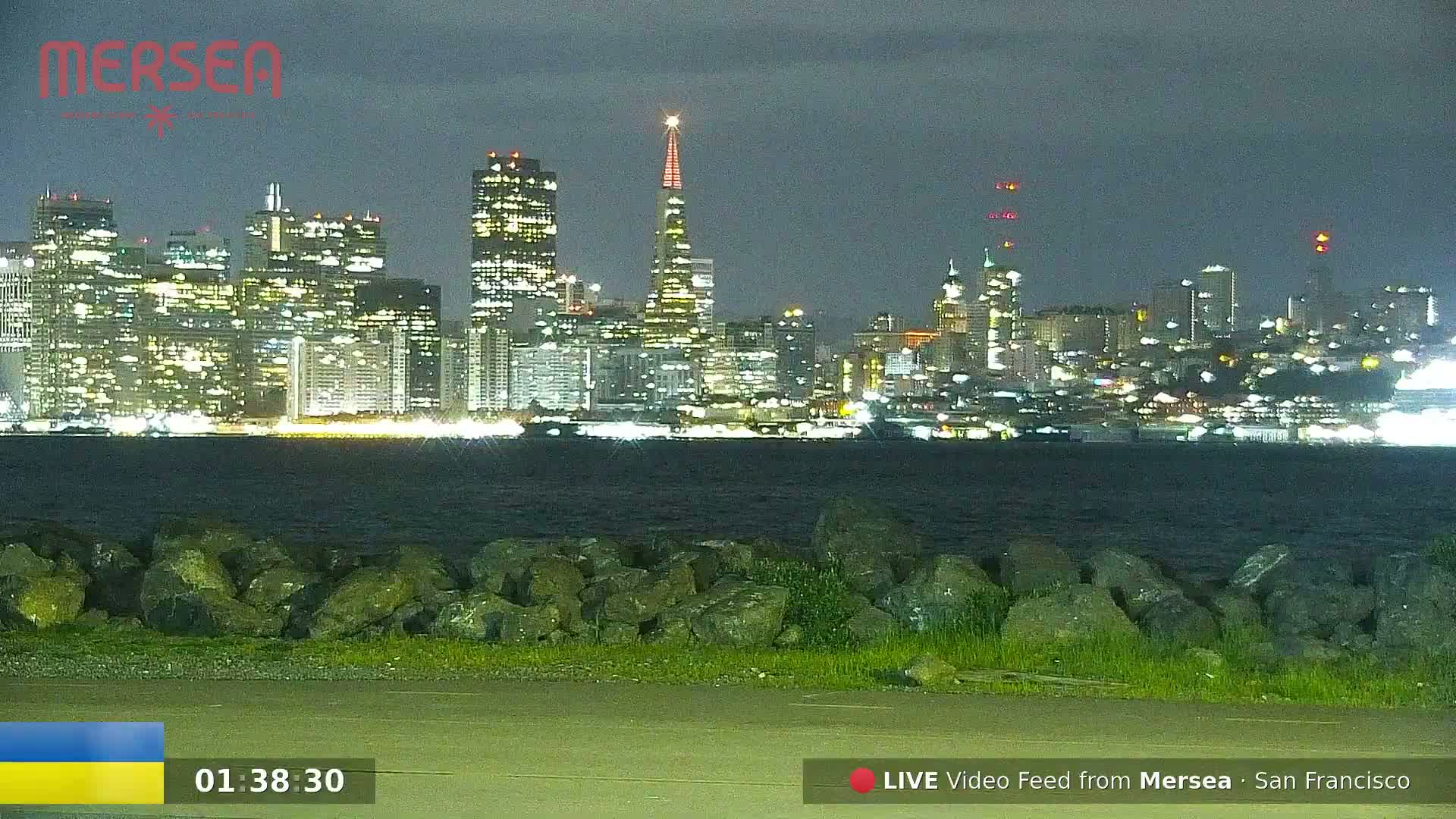 A nighttime view of a city skyline across a body of water, with a rocky shoreline in the foreground.