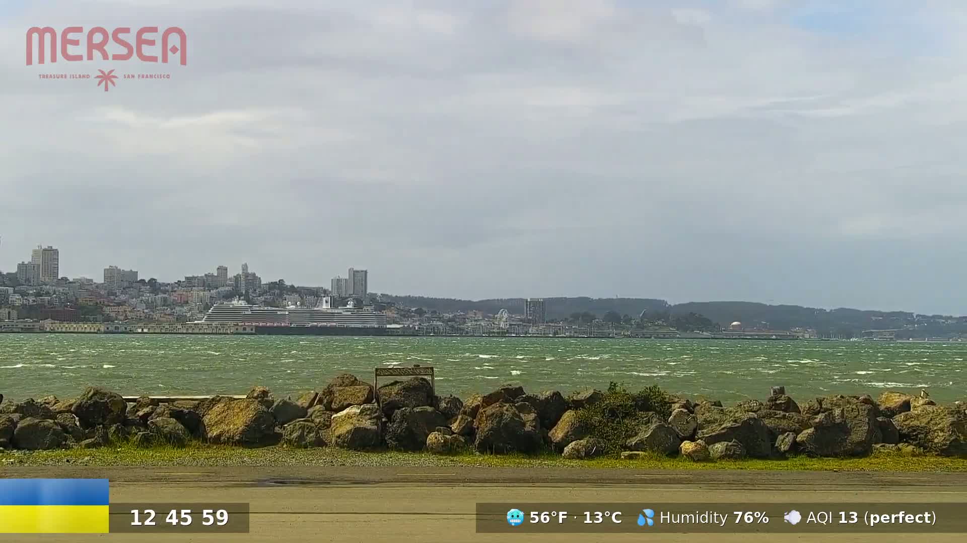 A large cruise ship is docked in a harbor, across a windy, green body of water from a rocky shoreline under a mostly cloudy sky.