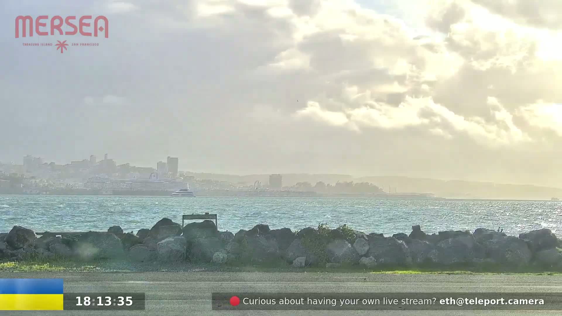 A partly cloudy day shows a body of water with a large cruise ship in the distance, a rocky shoreline in the foreground, and a paved area.