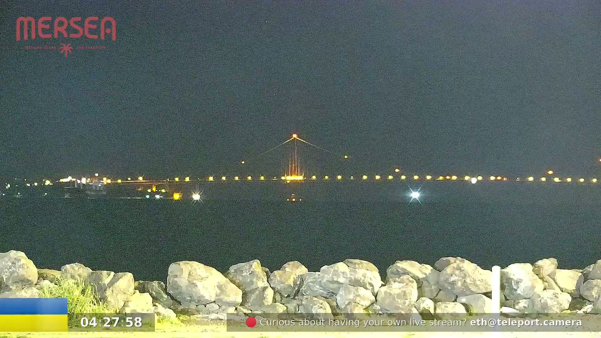 A nighttime view of a lit-up suspension bridge over dark water, seen from behind a low rock wall.