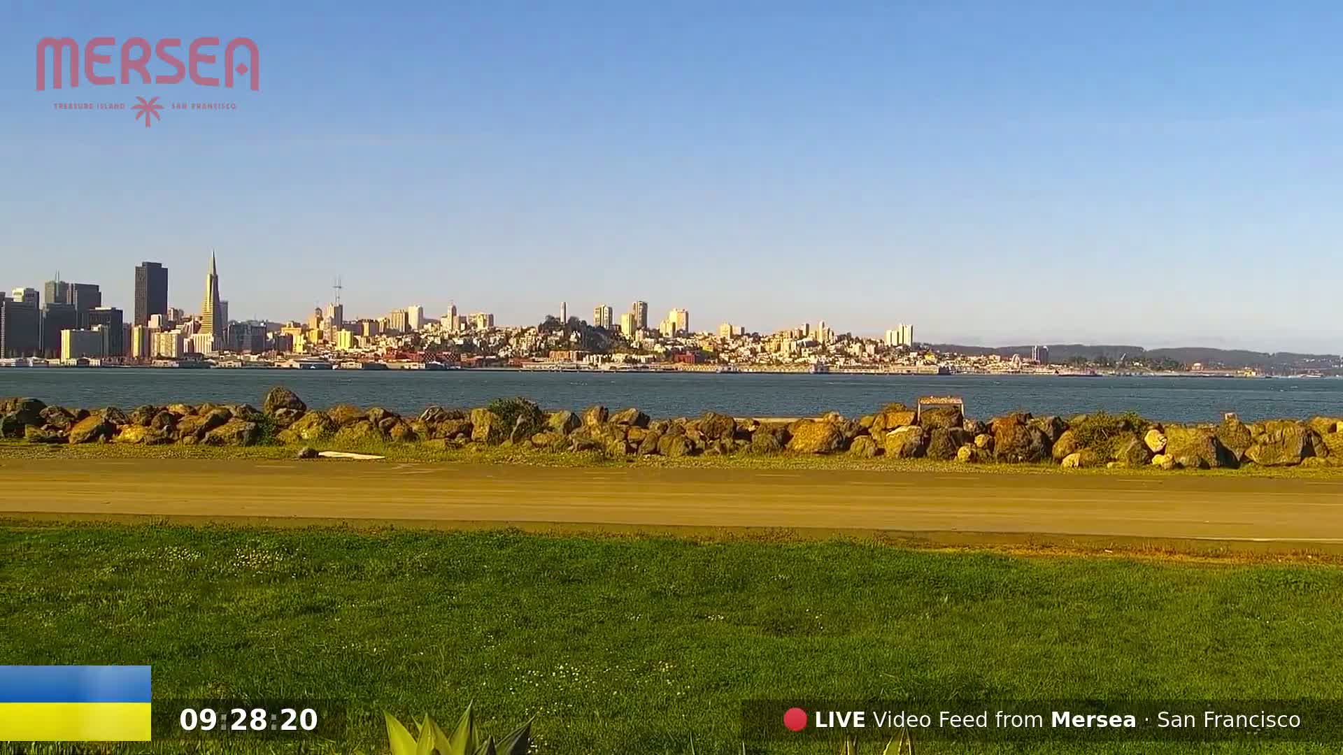 A grassy area with a paved path overlooks a calm body of water, beyond which the San Francisco skyline is visible under a clear, sunny sky.