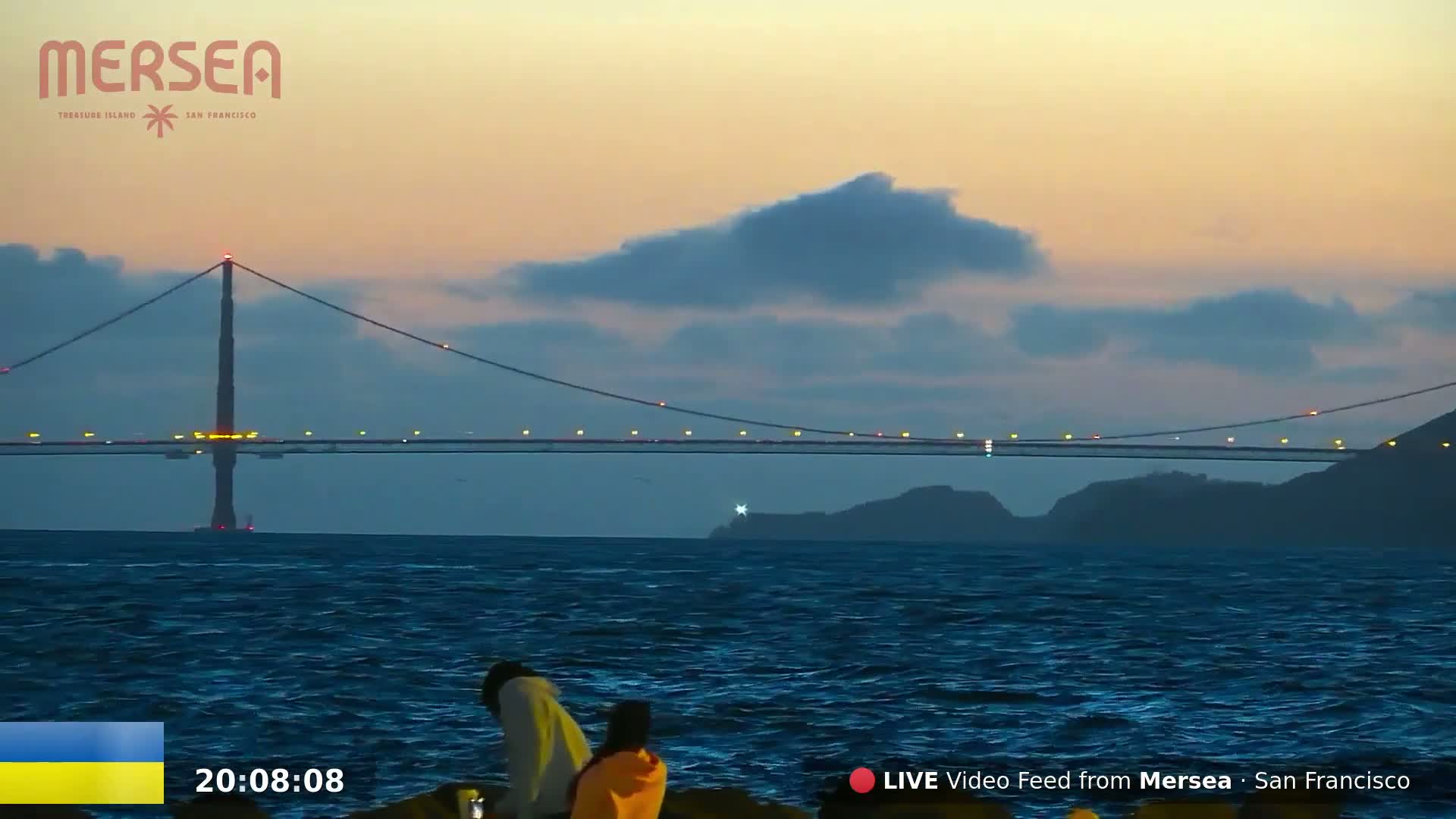 Two people sit on rocks overlooking a calm body of water at dusk, with a bridge illuminated with lights in the background under a partly cloudy sky.