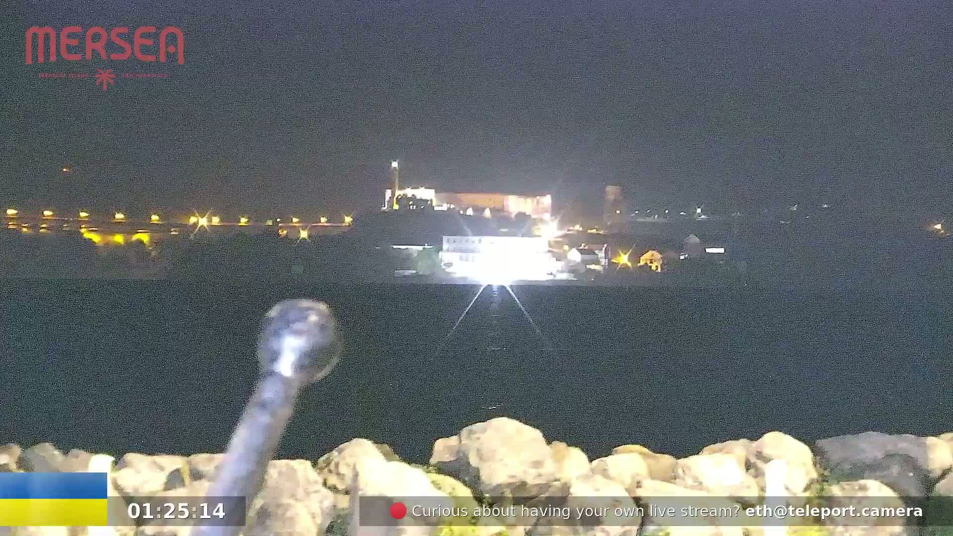A nighttime, low-angle view shows a body of water with a distant, illuminated town or city on the far shore.