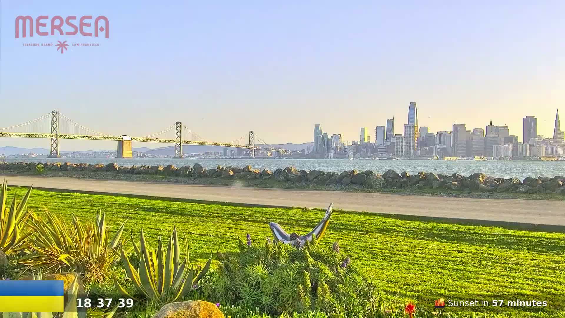 A grassy park area with a metal dolphin sculpture overlooks a calm body of water, a suspension bridge, and the San Francisco skyline under a clear, bright sky.