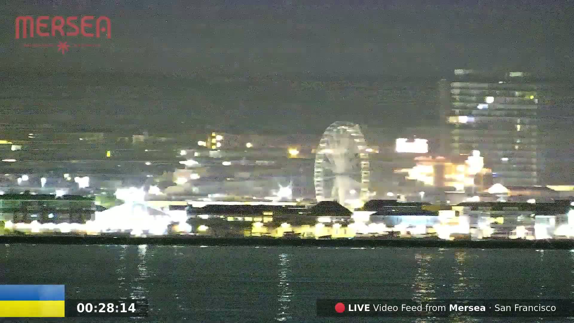 A nighttime view across dark water shows a brightly lit waterfront with a Ferris wheel and other buildings.