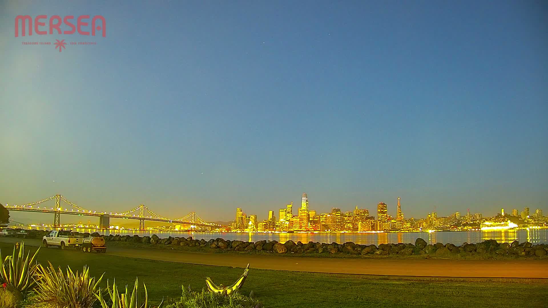 A nighttime view of a city skyline across a body of water, with a bridge illuminated in the distance under a clear twilight sky.