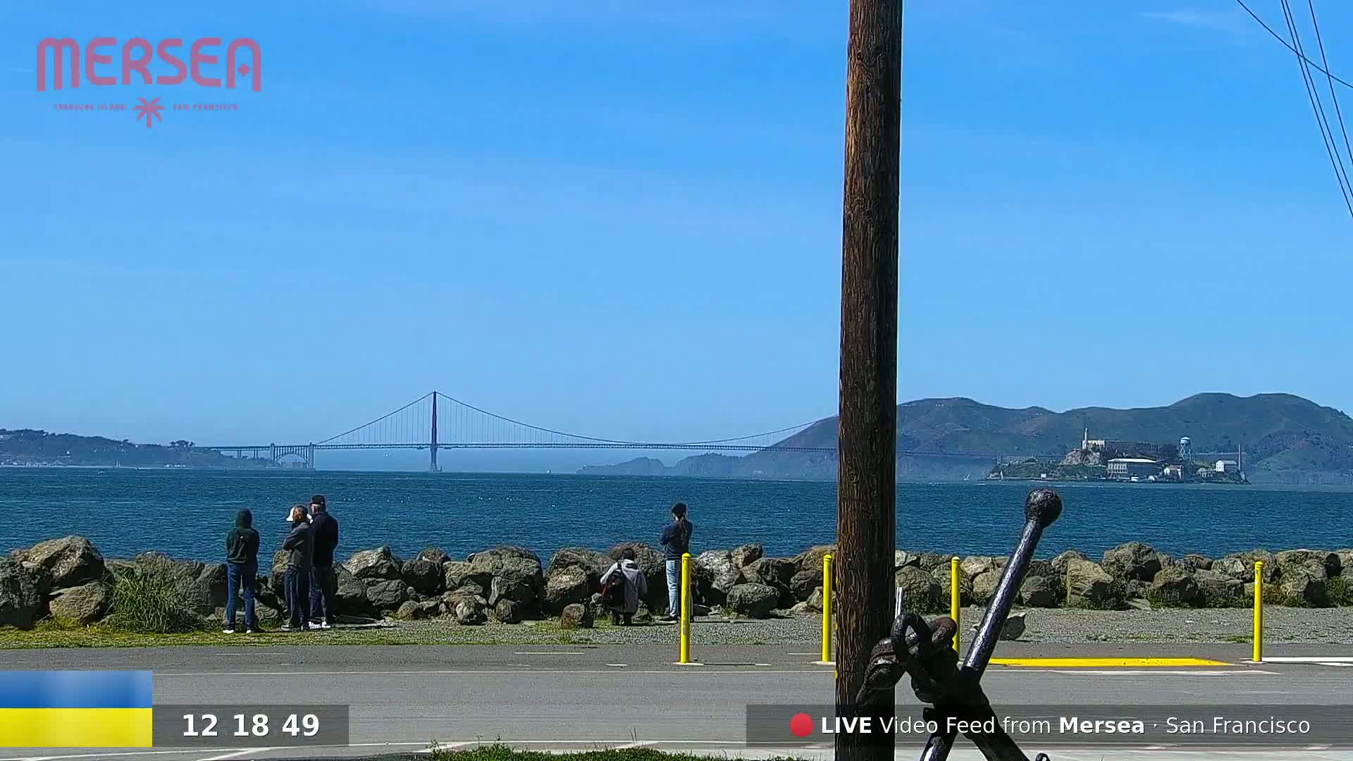A sunny day shows a group of people standing by a rocky shoreline overlooking a body of water with a bridge and Alcatraz Island in the distance.