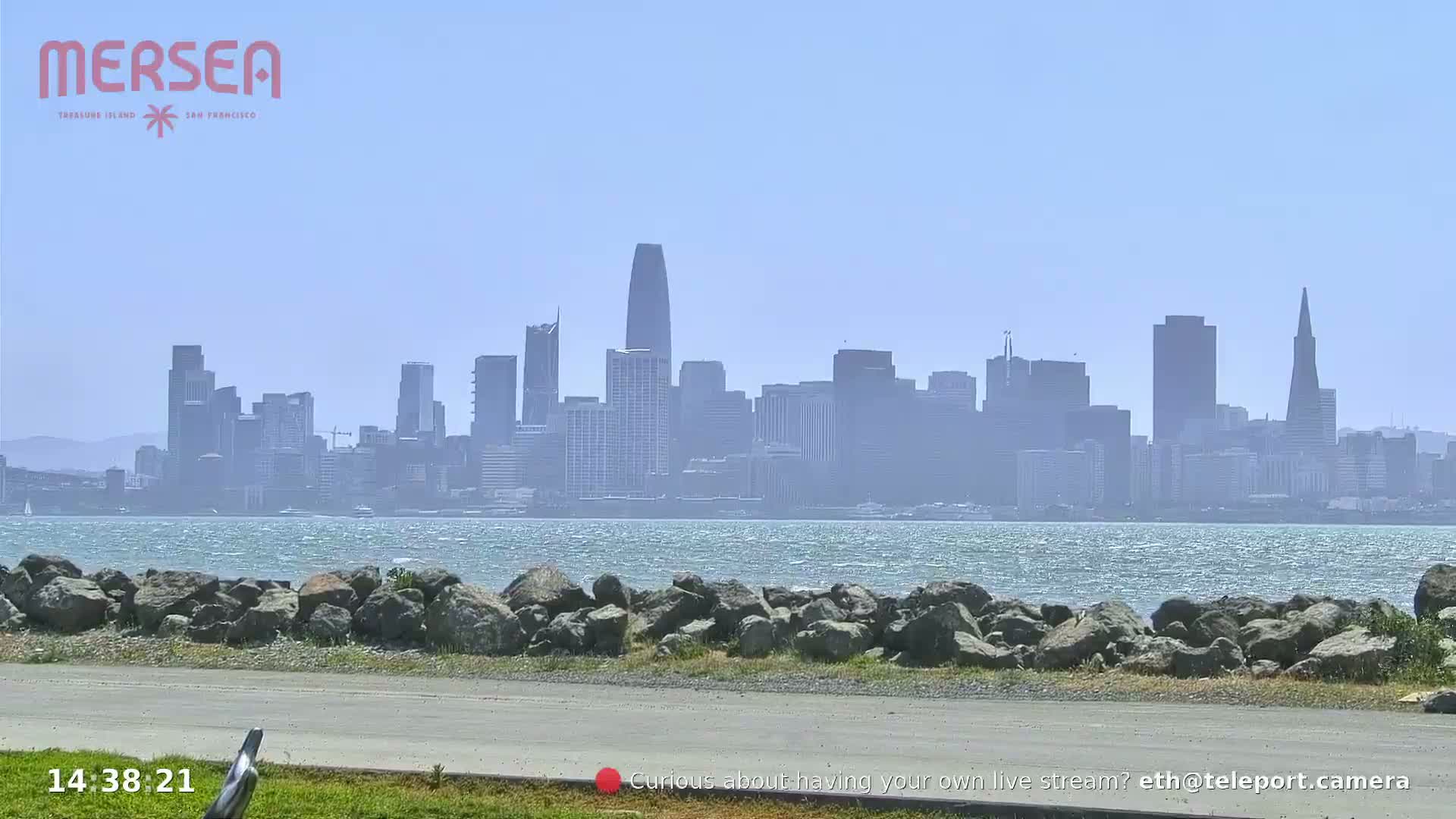 A hazy view of the San Francisco skyline across a body of water, with a rocky shoreline in the foreground.