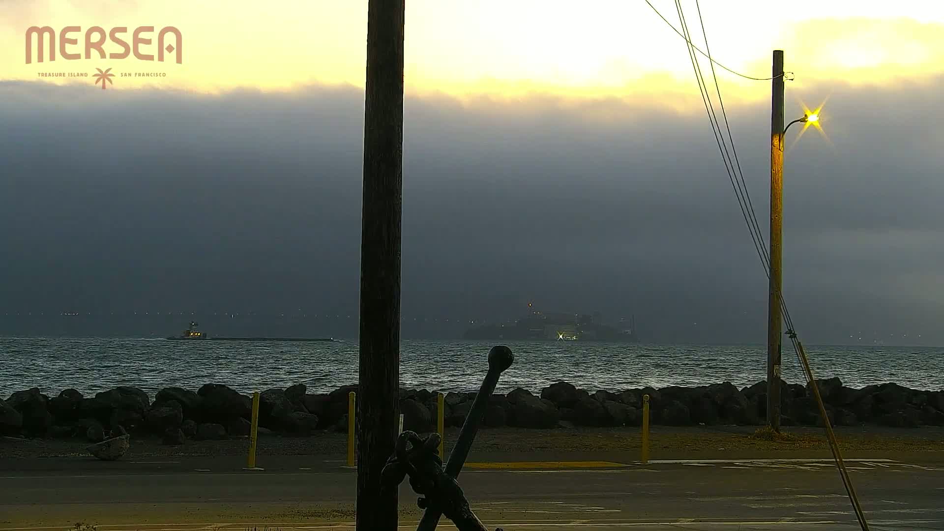 A partly cloudy, dusky evening scene shows a body of water with Alcatraz Island in the distance, a small boat, a rocky shore, and a large anchor in the foreground.