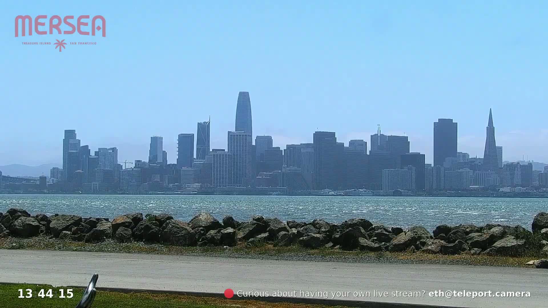 A city skyline is visible across a body of water on a sunny day, with a rocky shoreline in the foreground.