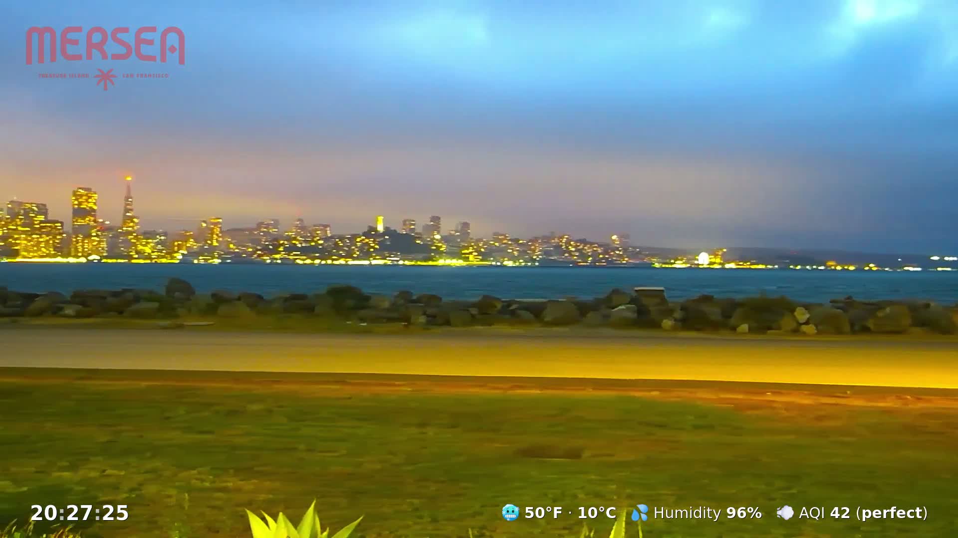A nighttime view of a city skyline across a body of water, under a mostly clear, dusky sky with high humidity.