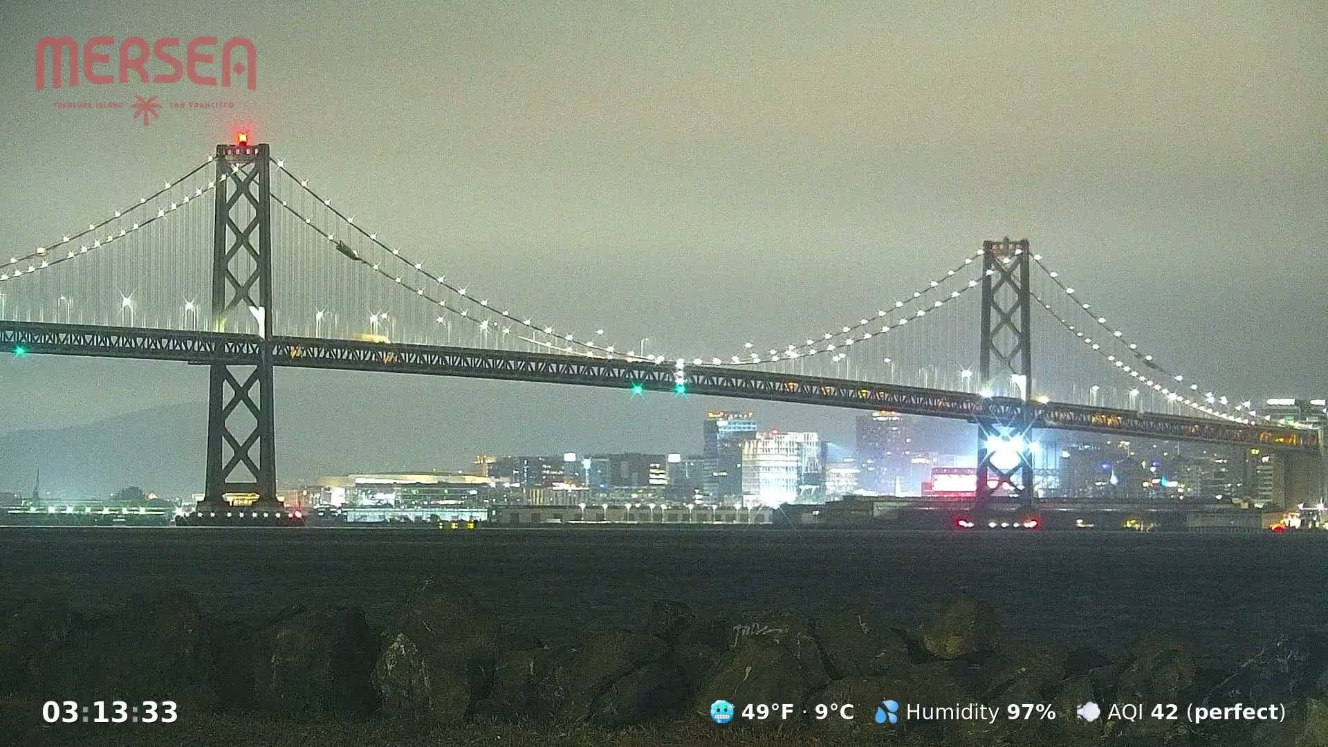 A suspension bridge illuminated at night is seen under a gray, overcast sky.