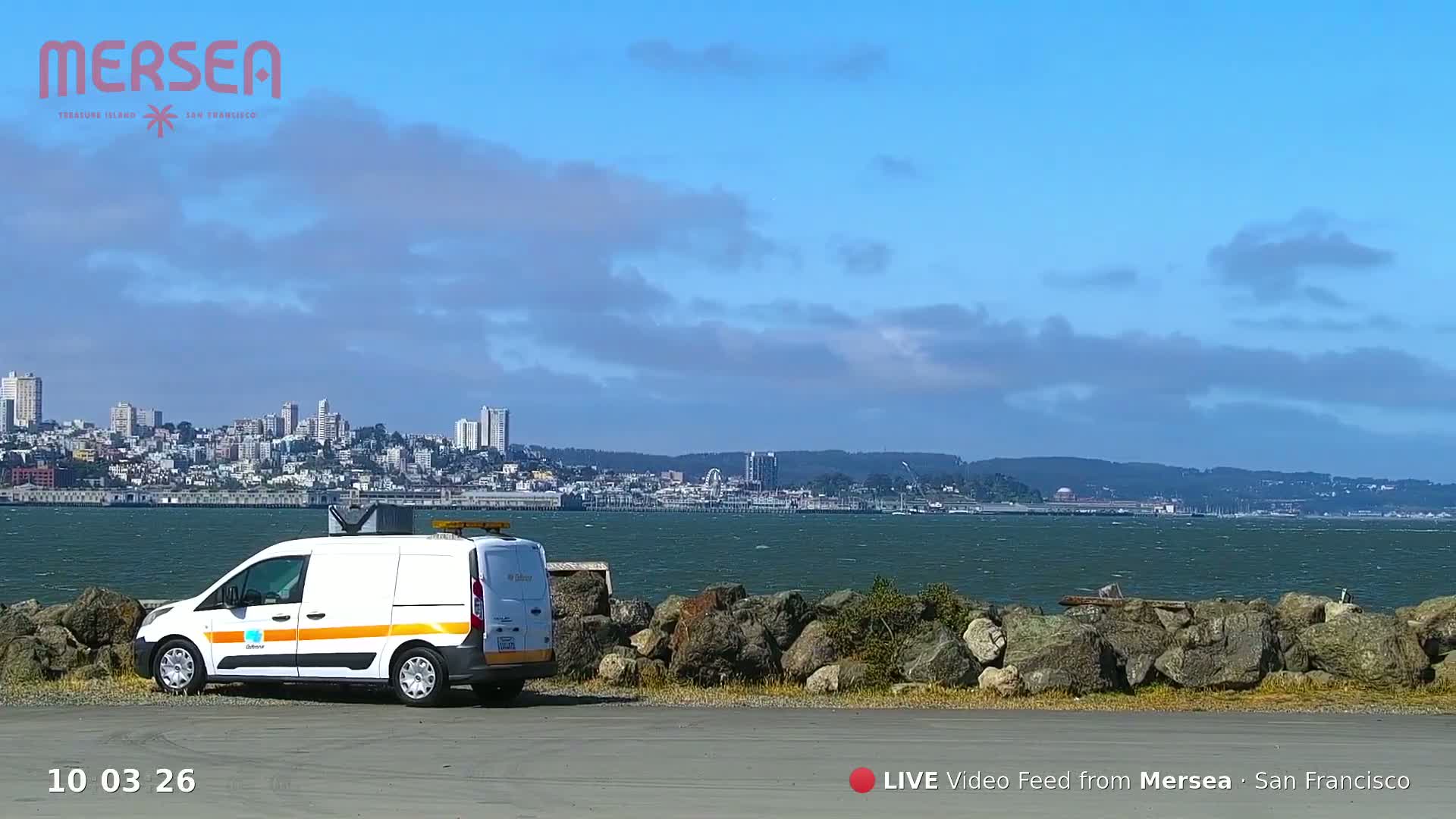 A white van is parked near a rocky shoreline, overlooking a city skyline across a body of water under a mostly sunny sky with some clouds.
