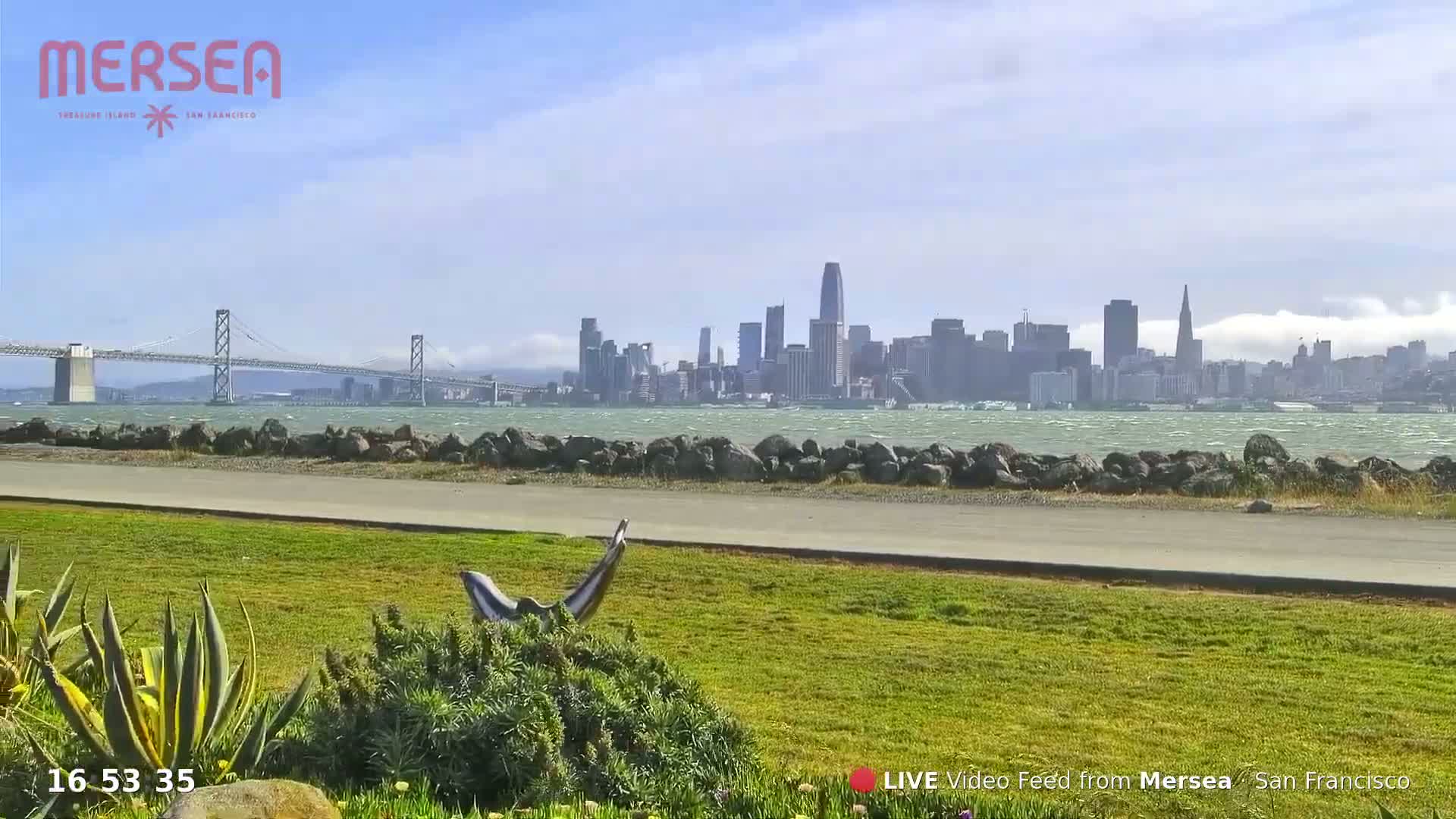 A grassy area with a sculpture overlooks a body of water, a bridge, and the San Francisco skyline under a partly sunny sky.