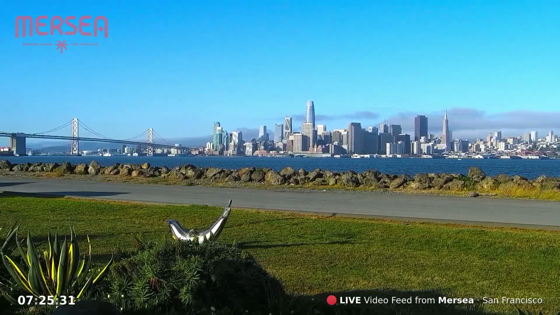 A clear, sunny day reveals a cityscape across a body of water, with a bridge in the foreground and a metallic sculpture on a grassy area.