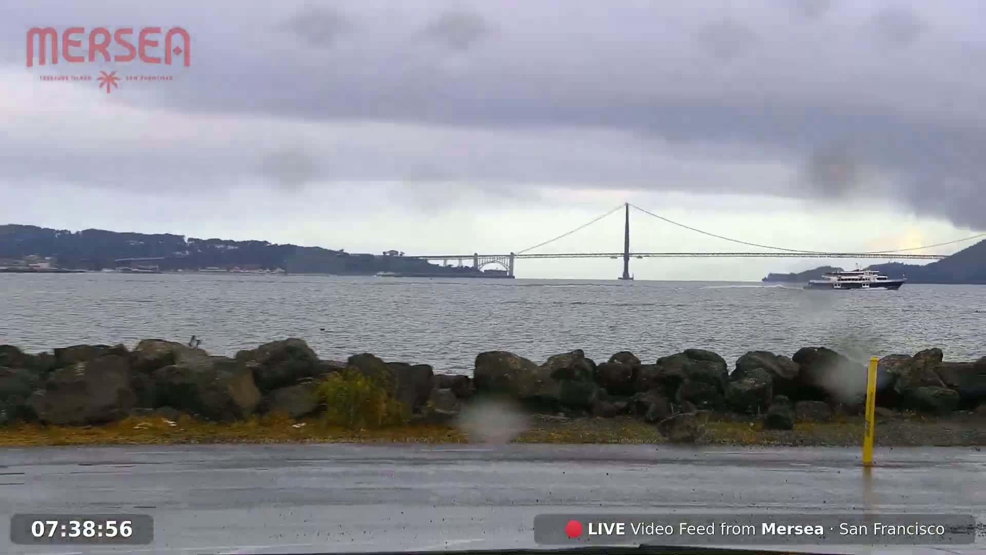 A ferry passes under a suspension bridge on a rainy day, with a rocky shoreline in the foreground.
