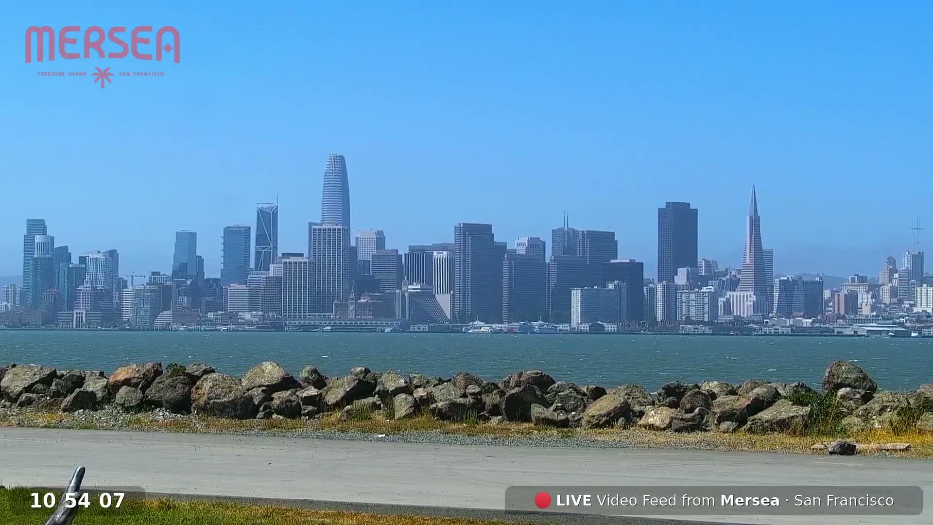 A sunny day reveals a cityscape across a body of water, viewed from a rocky shoreline.