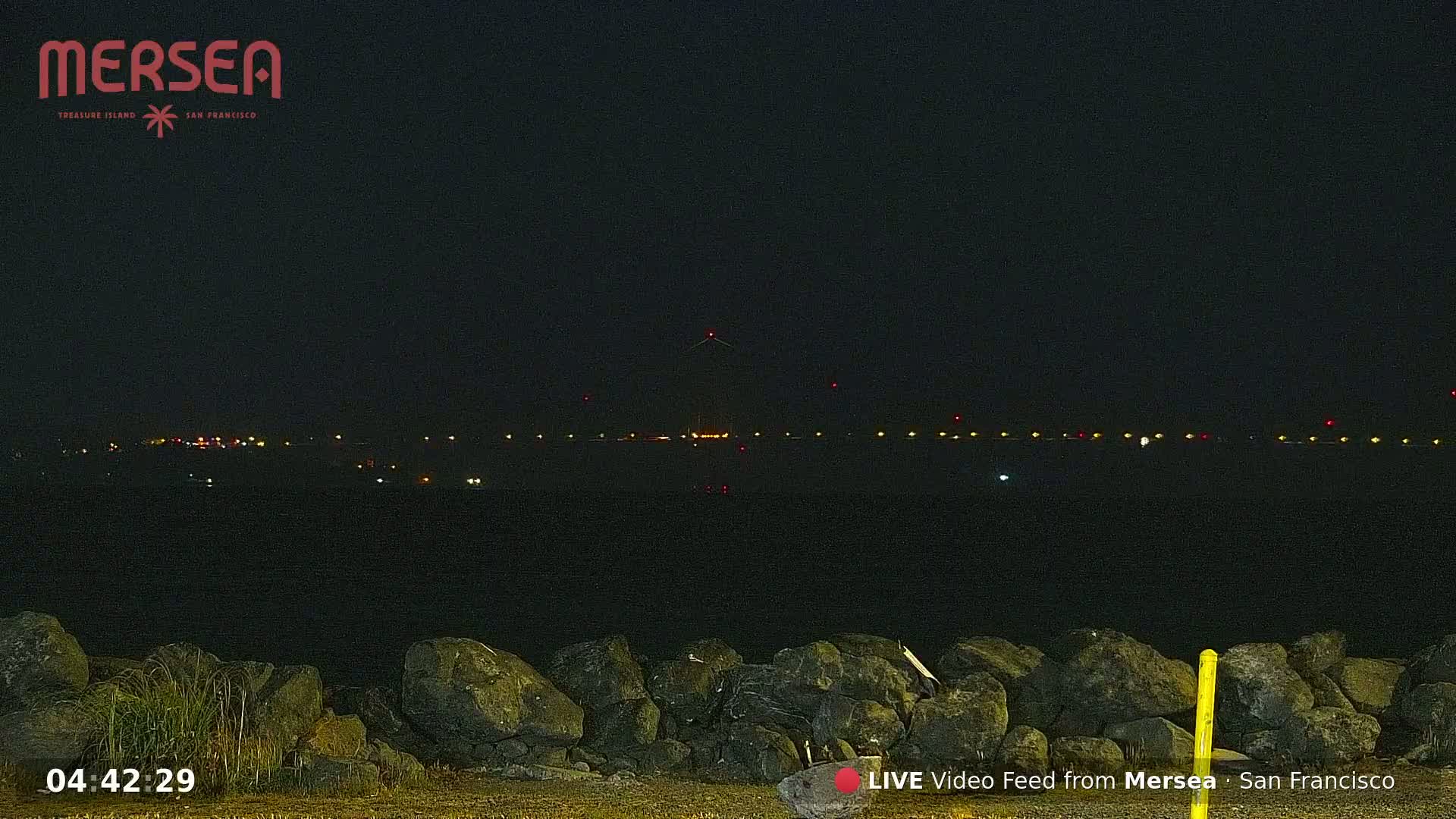A nighttime view of a distant, illuminated shoreline across a dark body of water, from behind a rocky embankment.