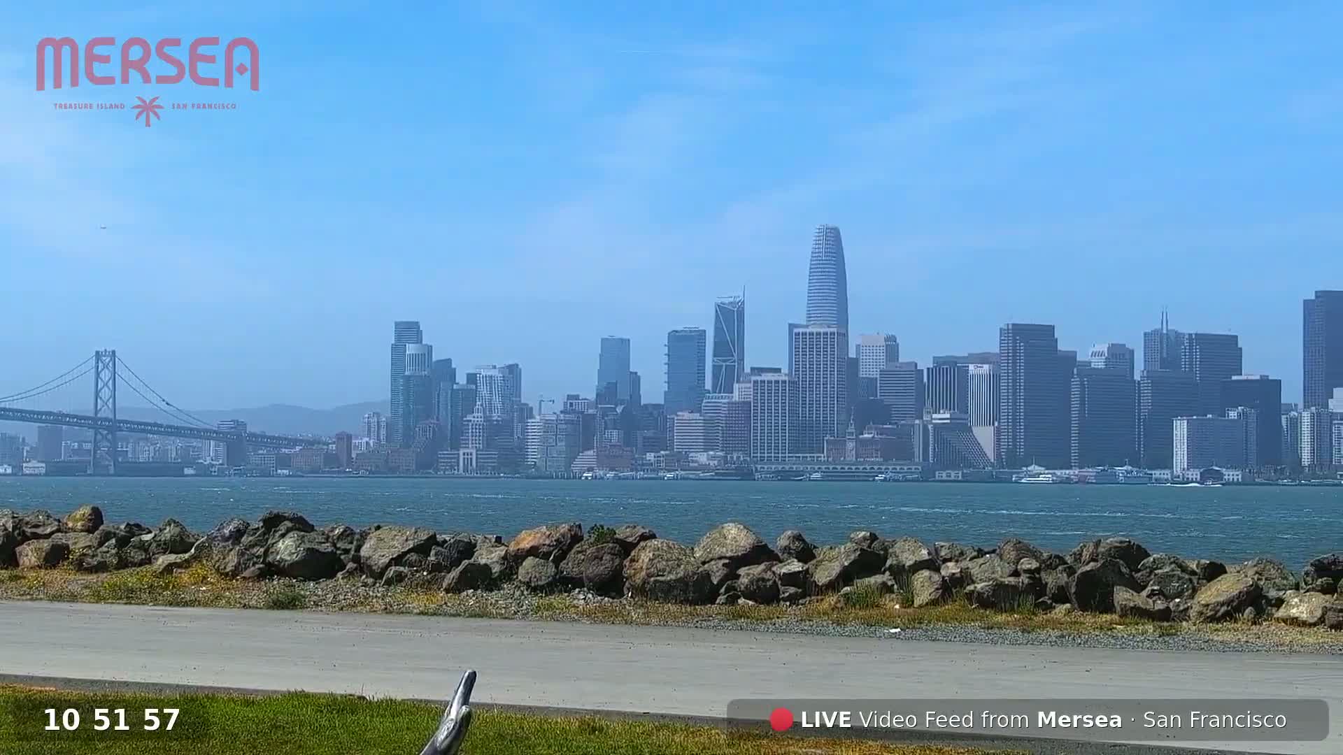 A city skyline is visible across a body of water on a clear, sunny day, with a bridge in the foreground.