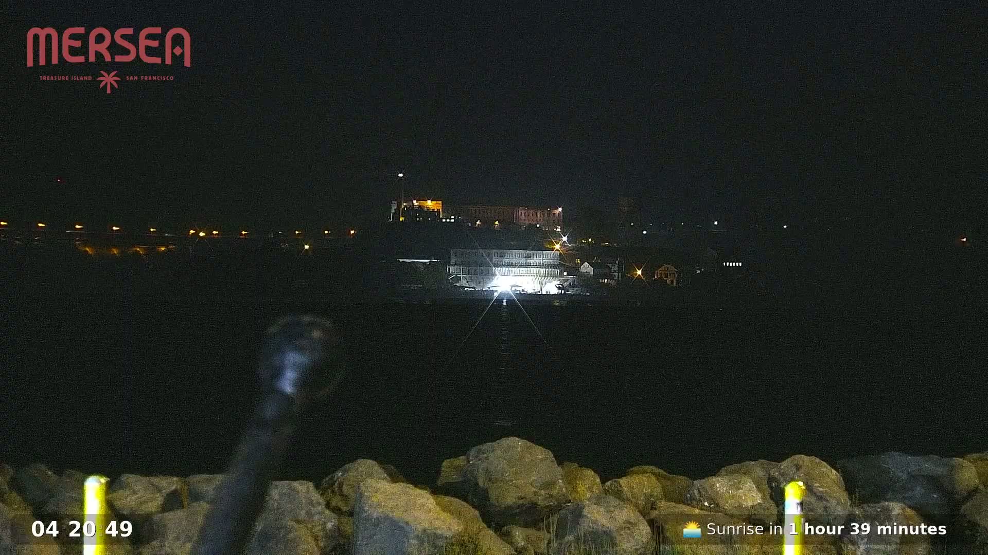 A nighttime view of a waterfront scene with a large building illuminated in the distance, partially obscured by darkness and viewed from behind a rocky embankment.