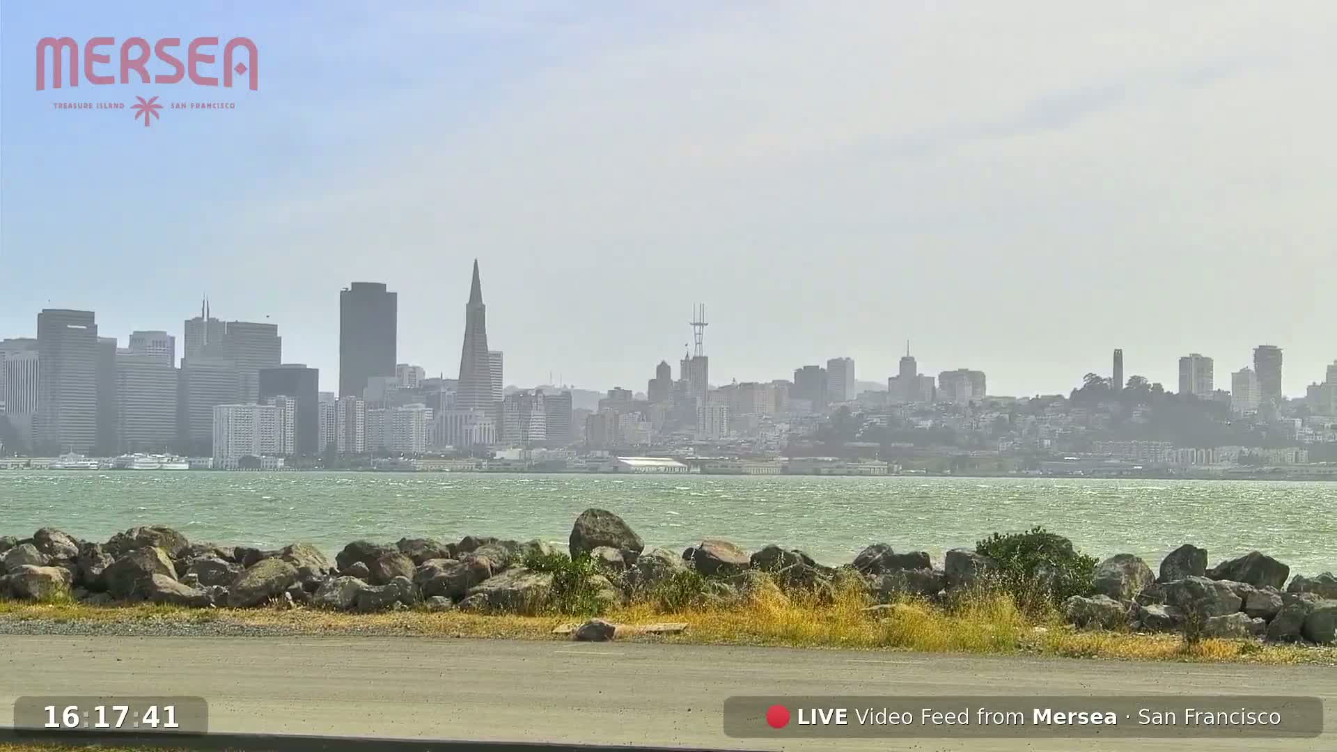 A hazy view of the San Francisco skyline across a body of water, with a rocky shoreline in the foreground.