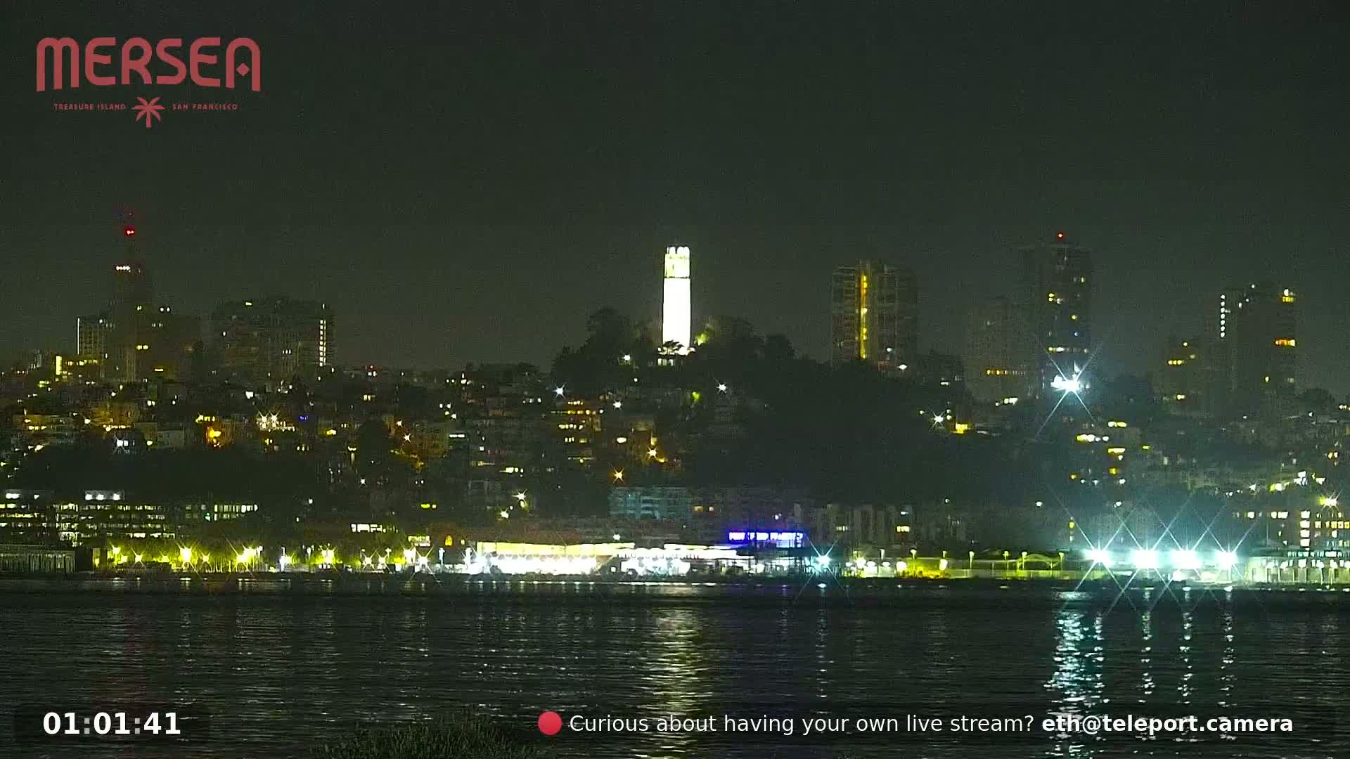 A nighttime cityscape with a brightly lit tower on a hill, reflected in calm water.