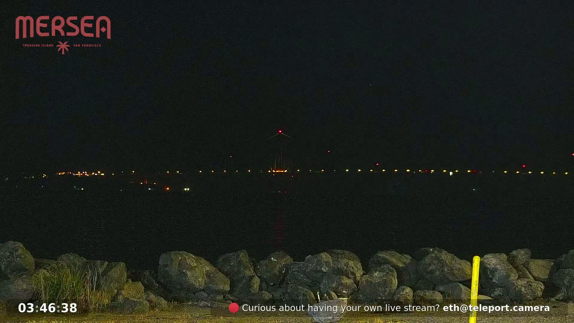 A nighttime view of a dark body of water with a distant line of lights and a tall structure with a red light at its top, viewed from behind a rocky shoreline.