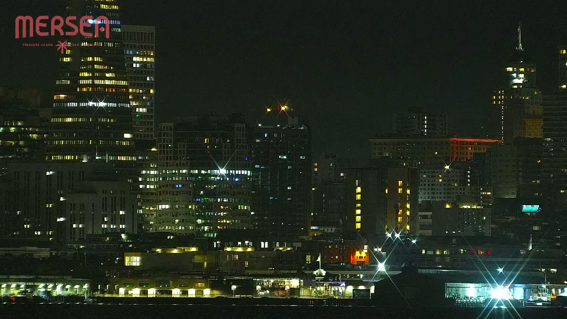 A nighttime cityscape of illuminated buildings along a waterfront, under a dark sky.