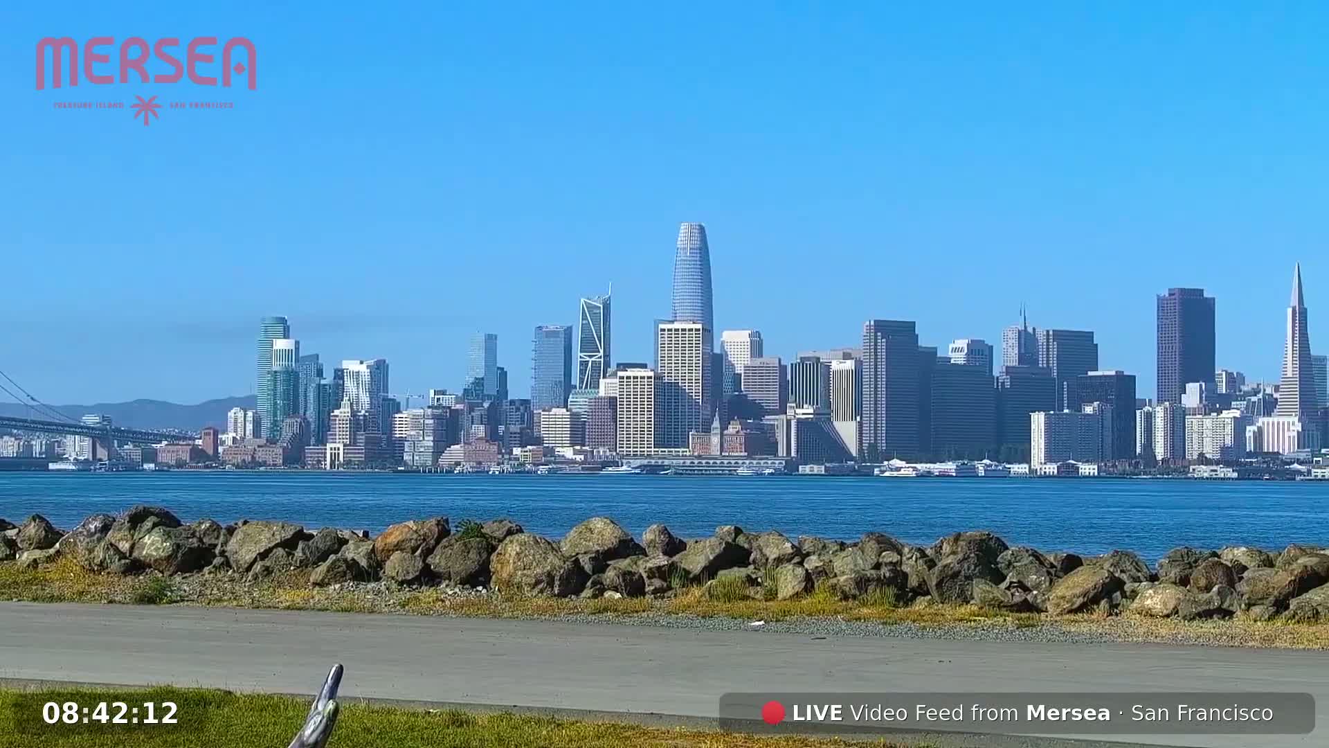 A clear, sunny day reveals a city skyline across a calm body of water, viewed from a rocky shoreline.