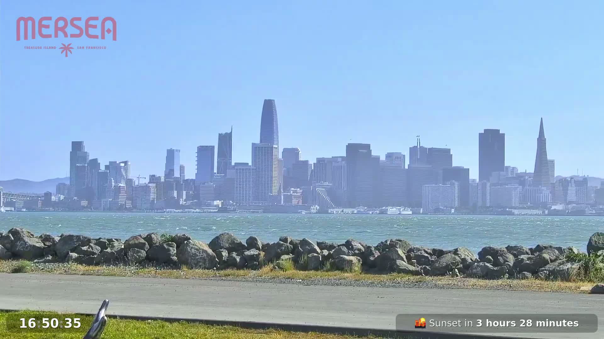 A hazy, sunny day shows a city skyline across a body of water with small waves, viewed from a rocky shoreline with a paved path.