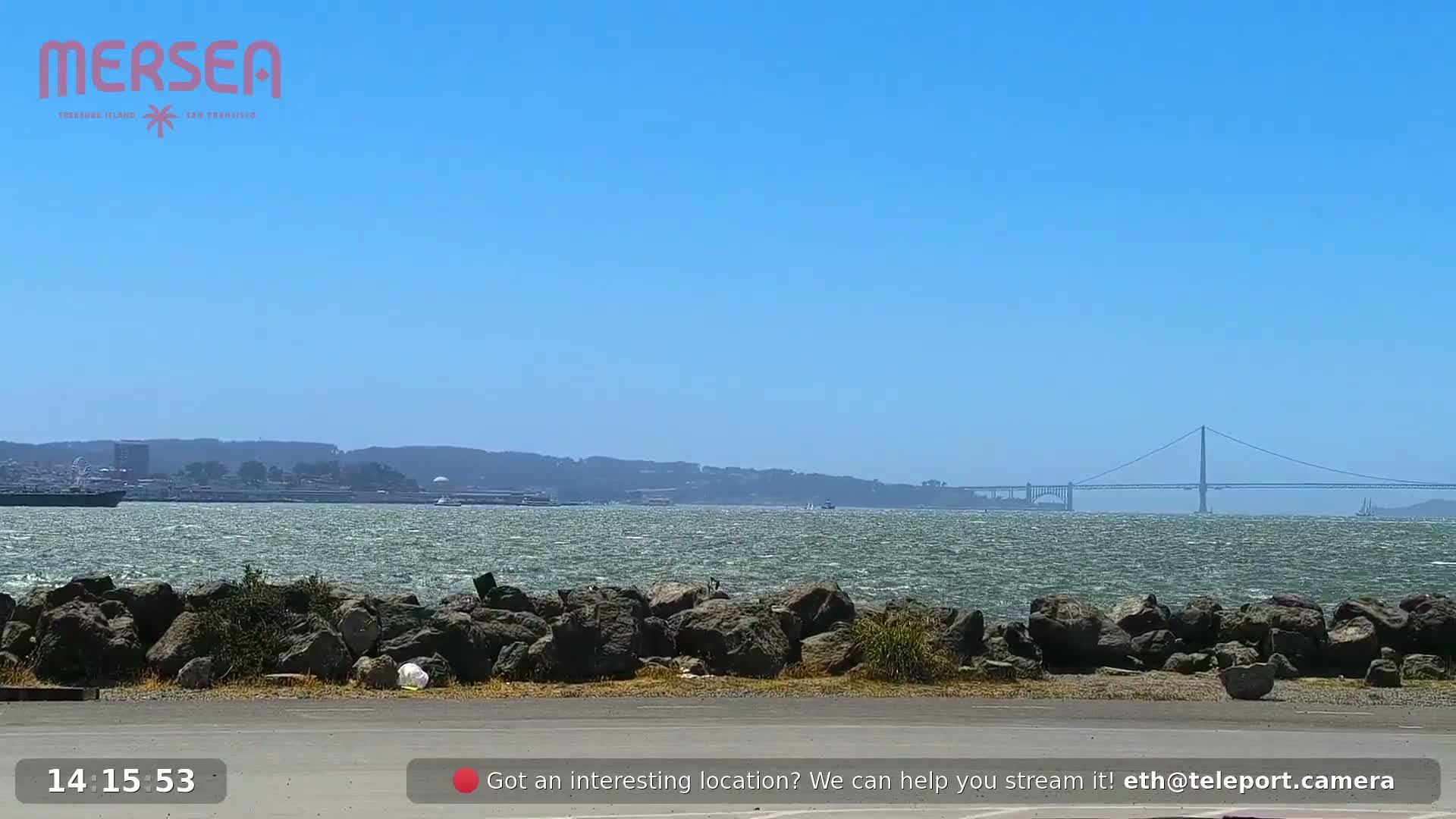 A rocky shoreline with a body of water in the foreground, showing a distant cityscape and a suspension bridge under a clear blue sky with a slight chop on the water.
